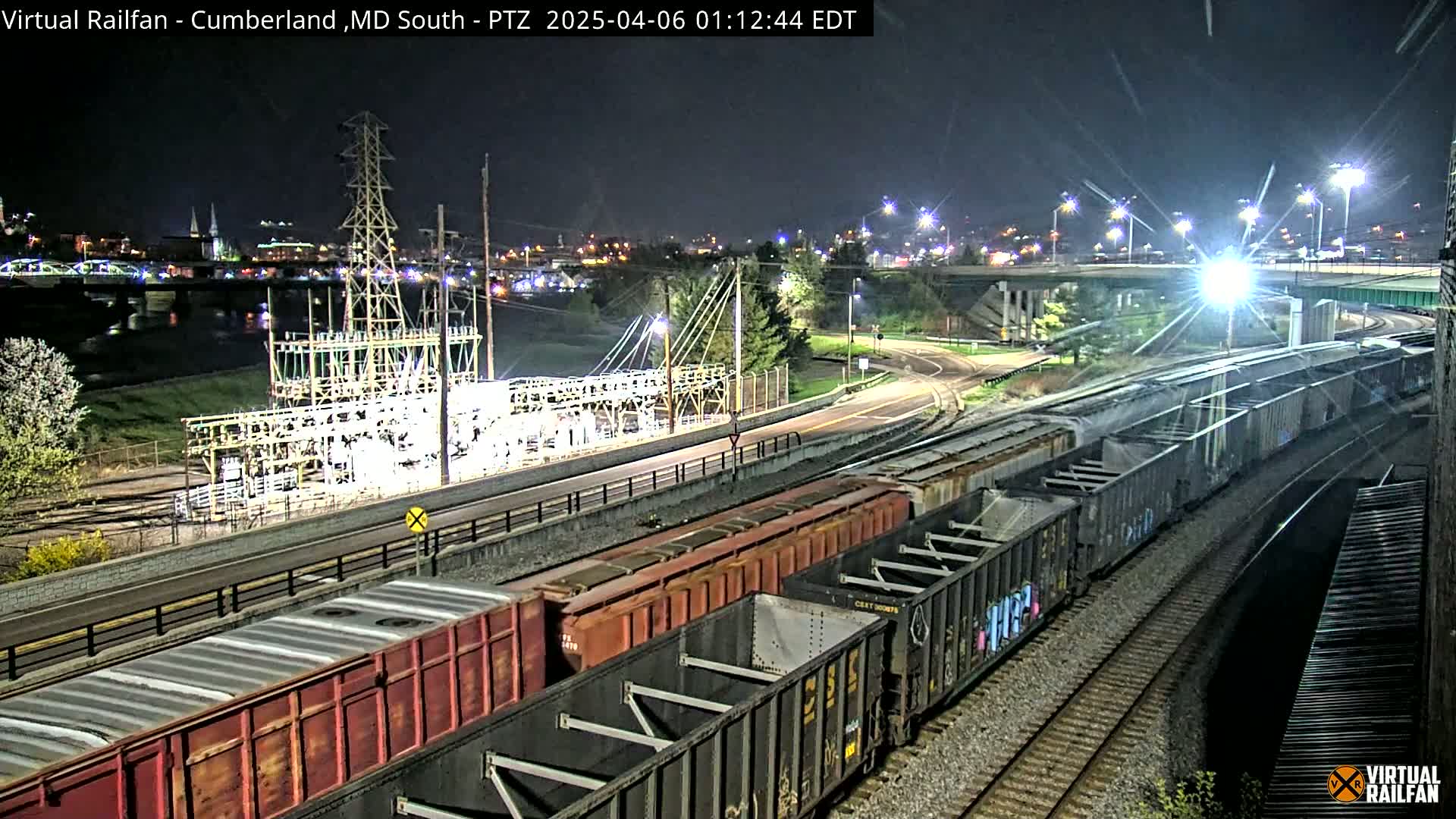 At night, a long freight train is passing by an electrical substation near a river and highway overpass under bright lights.