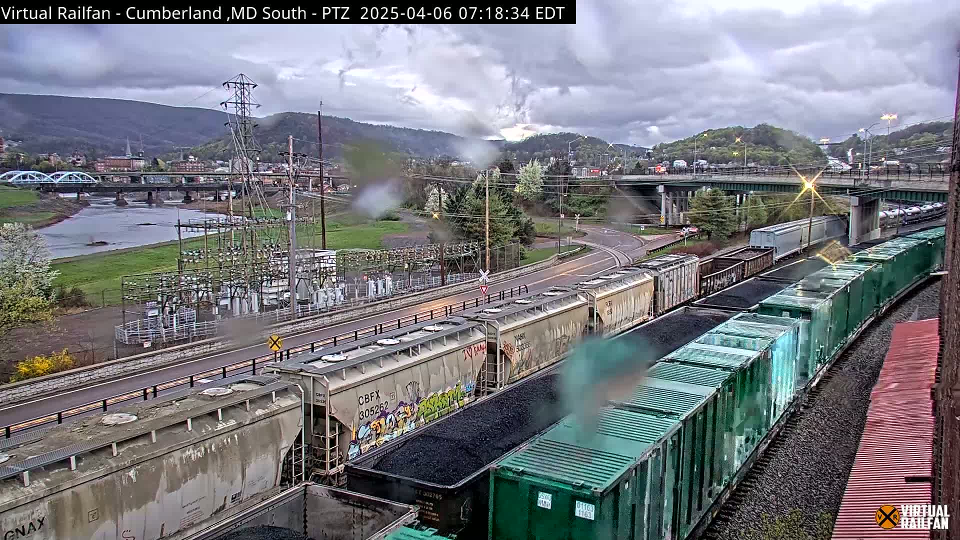 A long freight train carrying coal sits on tracks next to a river and highway under a cloudy, overcast sky.