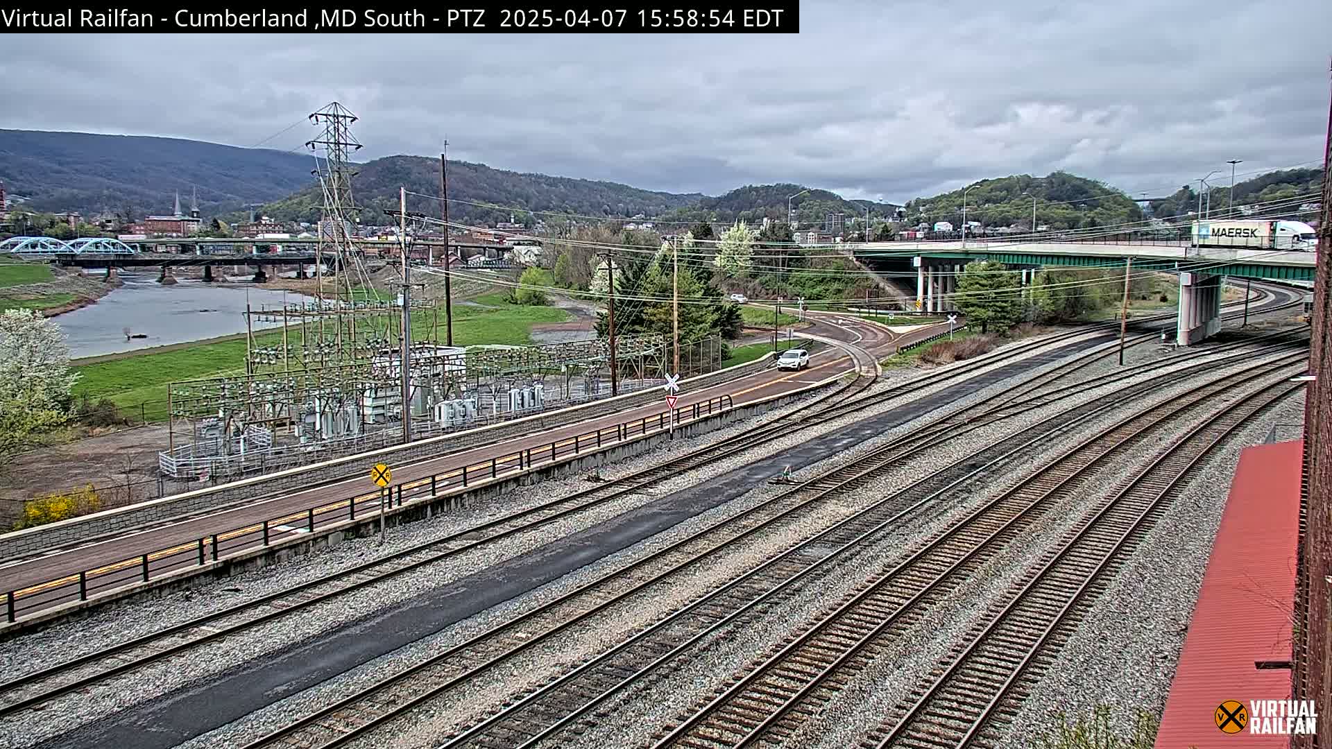 An overcast day reveals a view of multiple train tracks winding through a valley alongside a river, a bridge, and a power substation, with a highway overpass in the background.