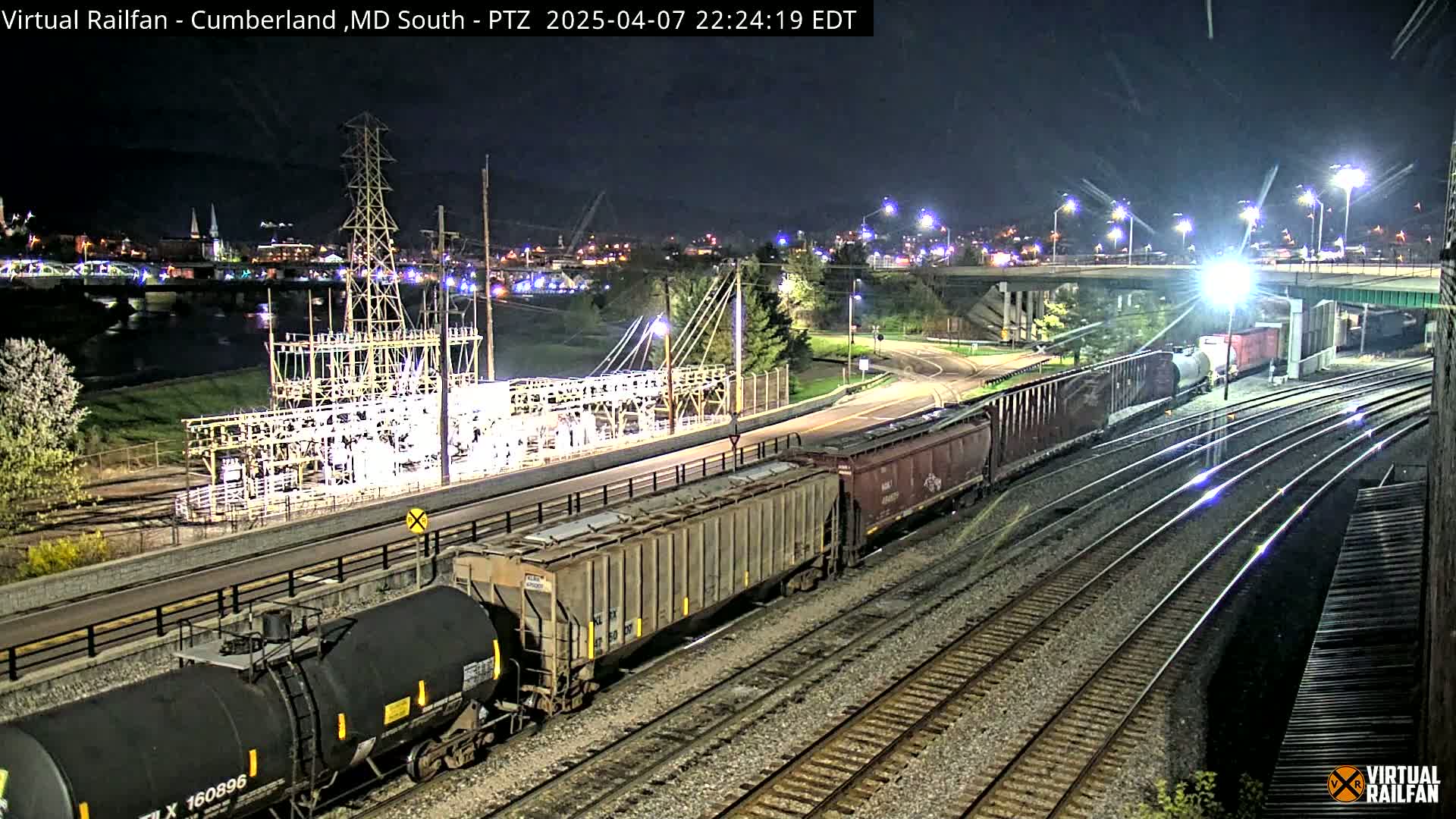 At night, a long freight train is seen moving along railroad tracks near a river and city lights, under a clear sky.