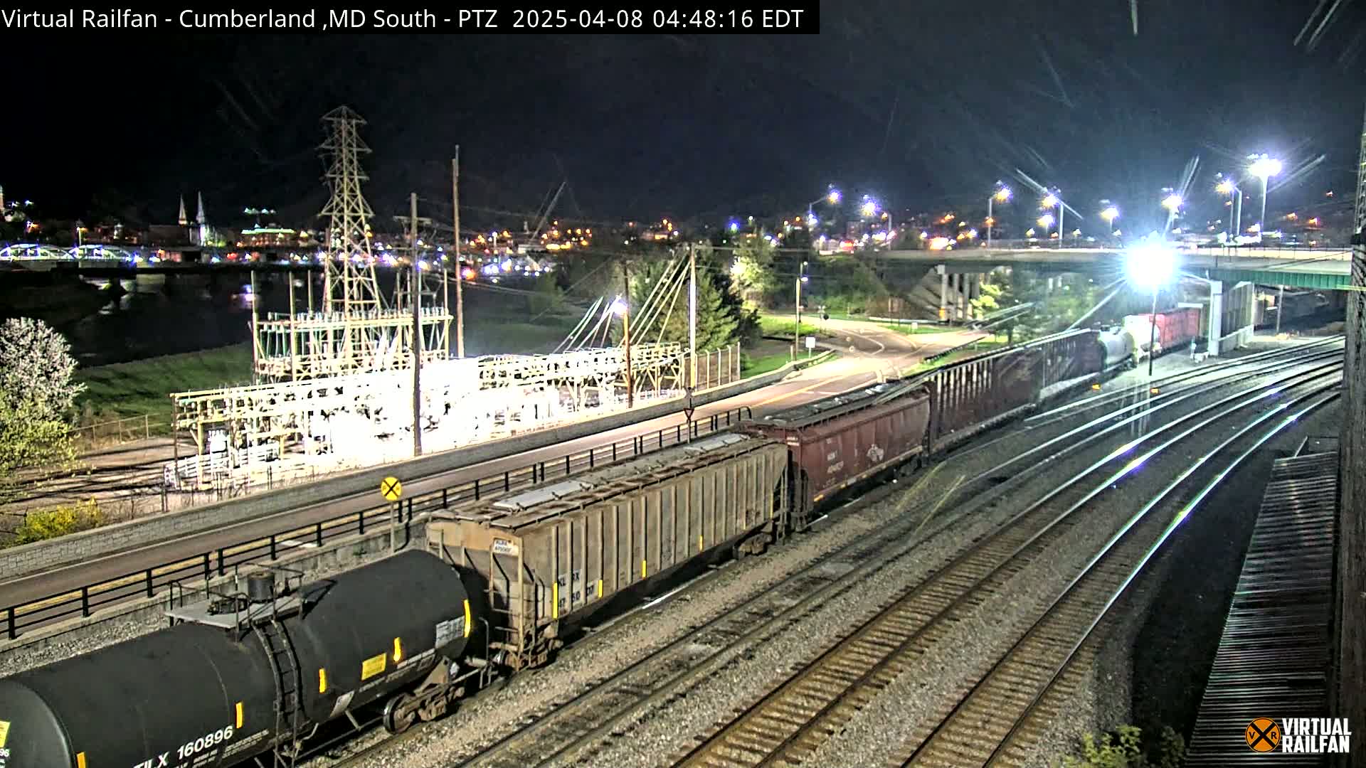 At night, a long freight train moves along several parallel railroad tracks near a power substation and a river, under bright lights.