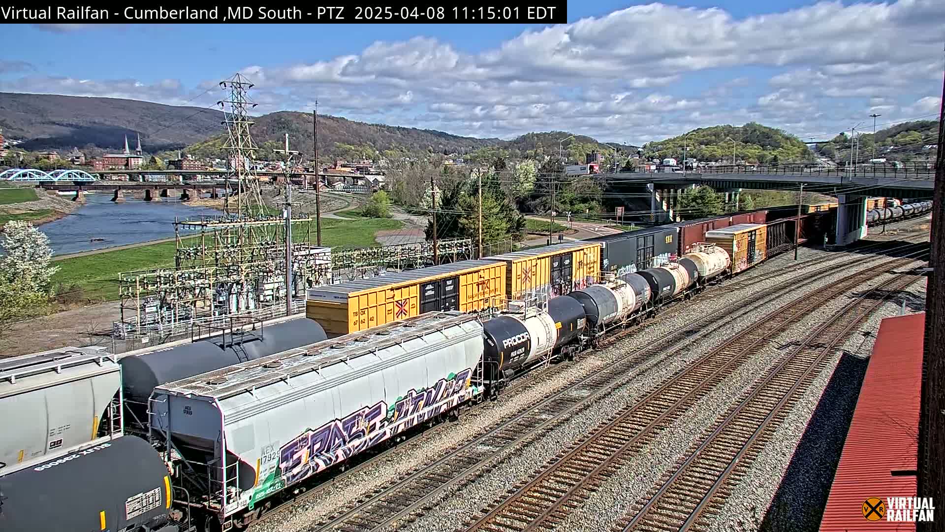A long freight train with various types of cars is on a set of railway tracks under a partly cloudy sky, next to a river and a town nestled in a valley.
