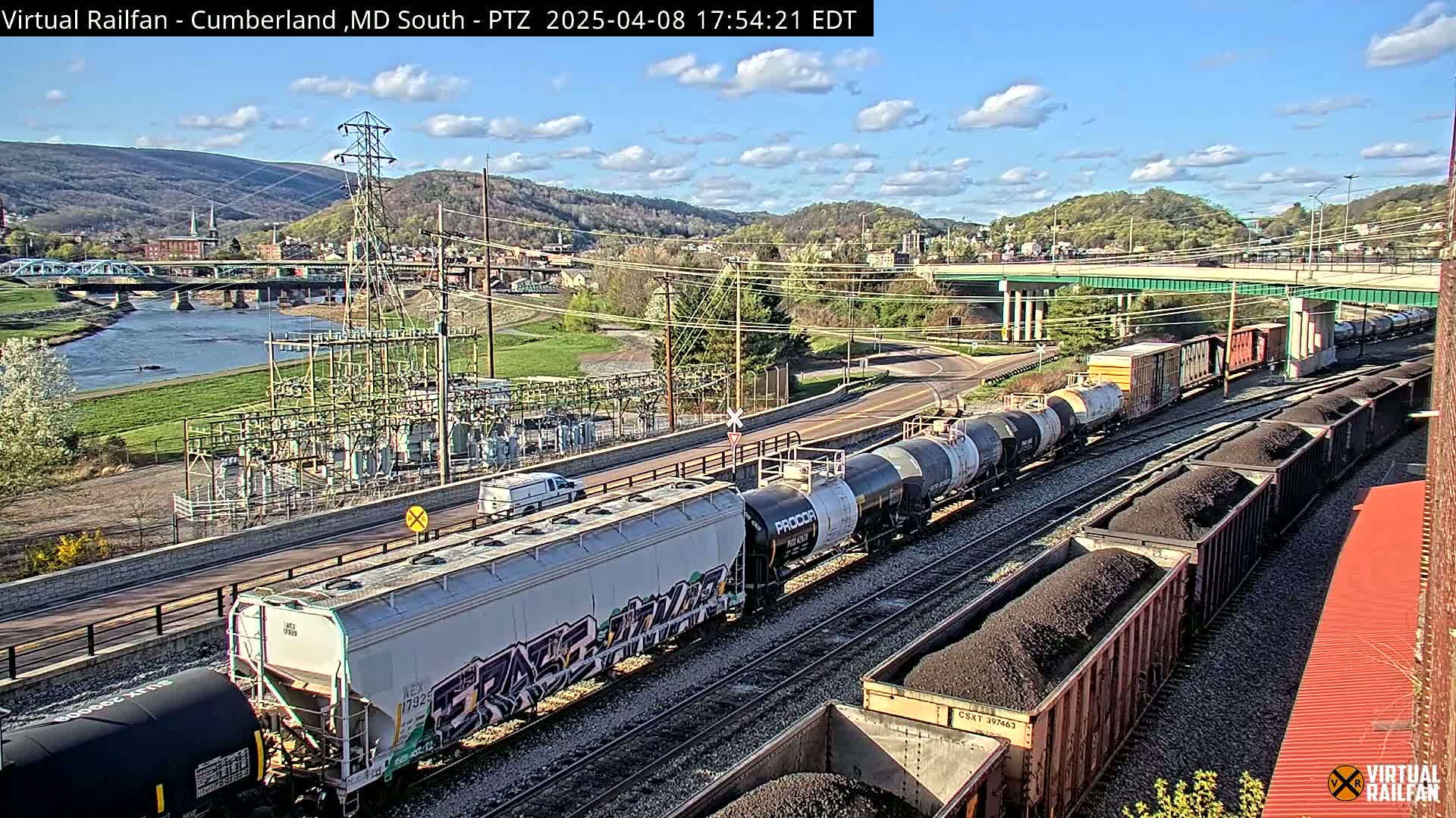 Under a partly cloudy sky, a long freight train carrying various cargo passes by a river and a town, with a highway overpass in the background.