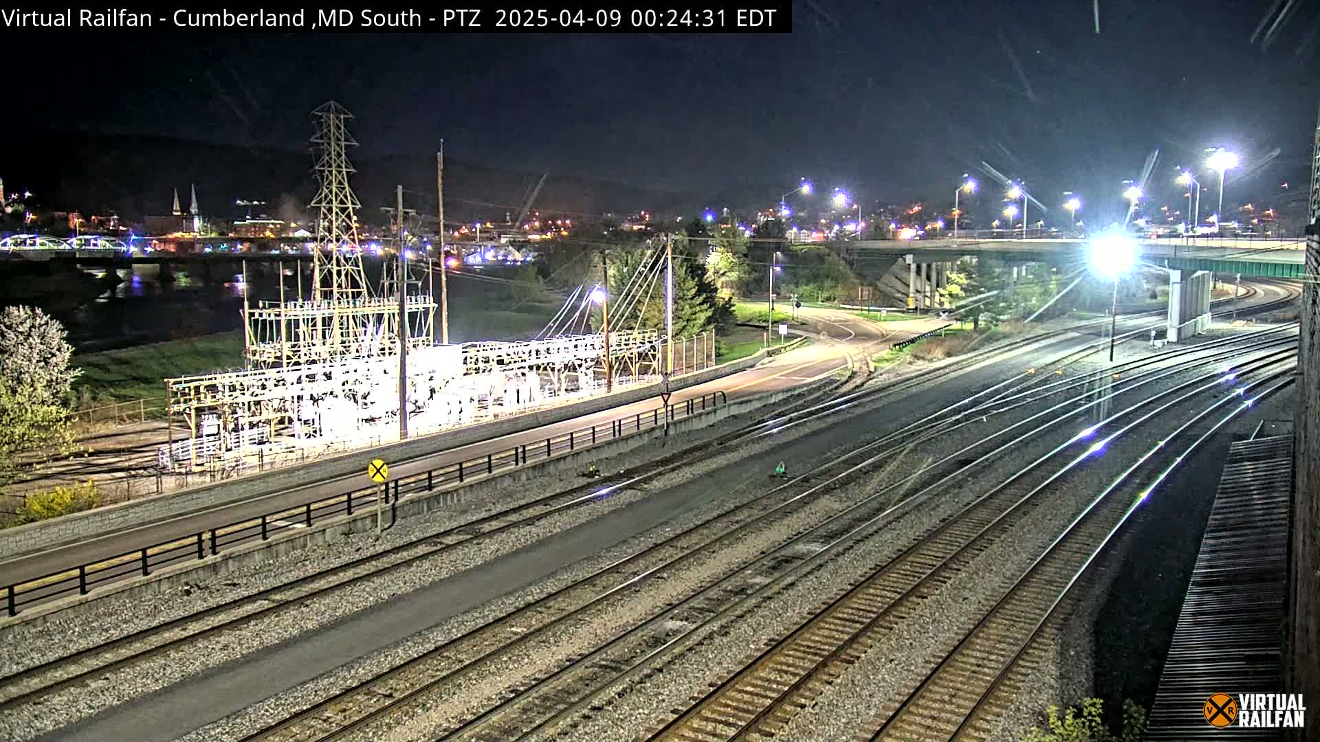 A nighttime view of railroad tracks and an electrical substation near a river, with a city skyline in the background under clear skies.