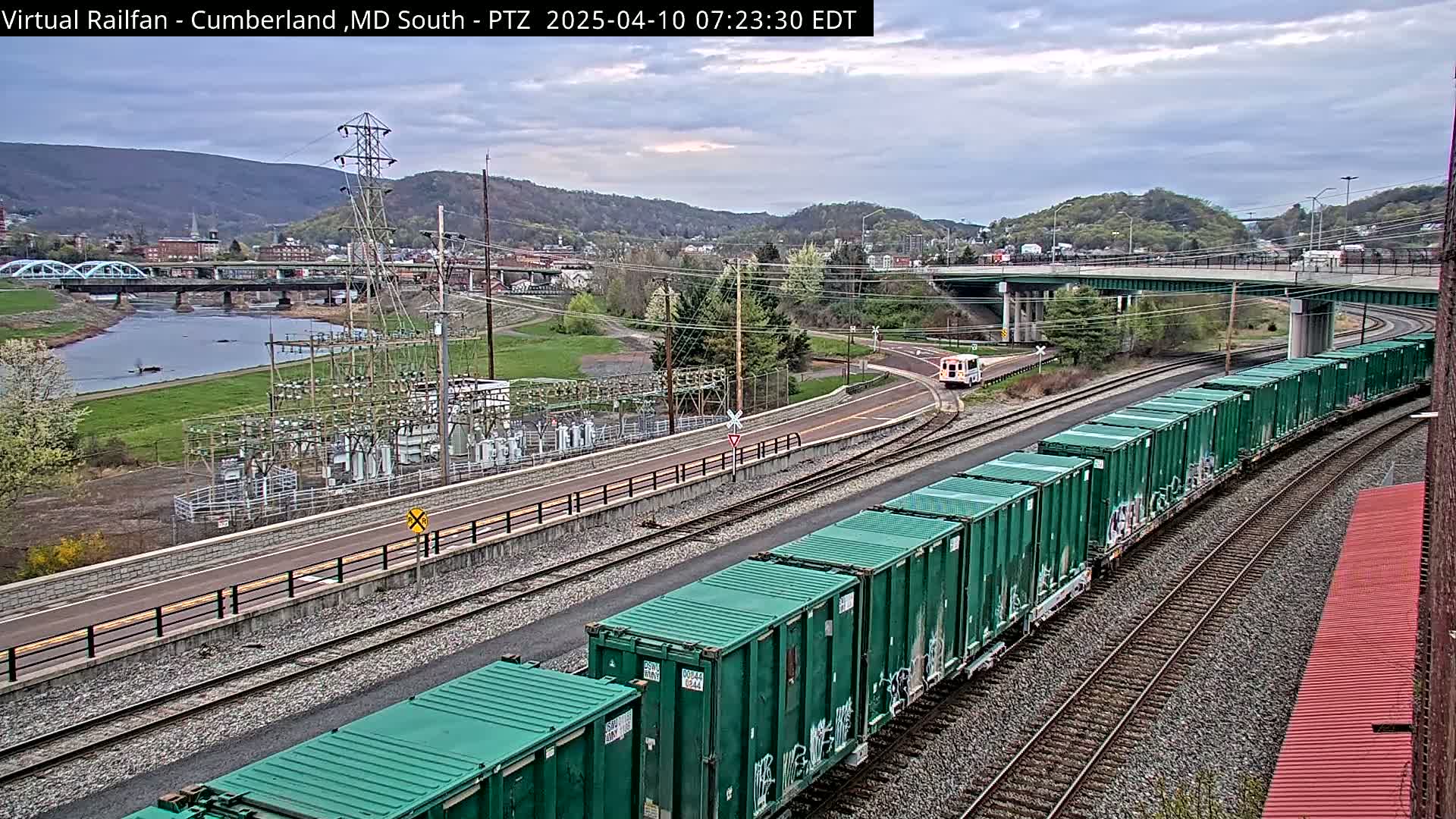 A long freight train with green containers curves along railroad tracks near a river, under an overcast sky, with a small town and hills in the background.