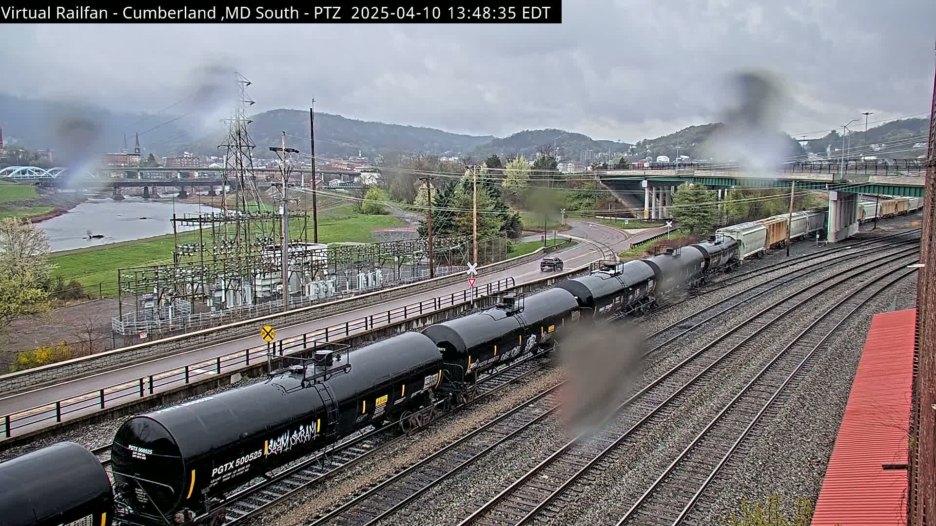A long freight train of black tanker cars is seen on a railroad track next to a river under an overcast sky.