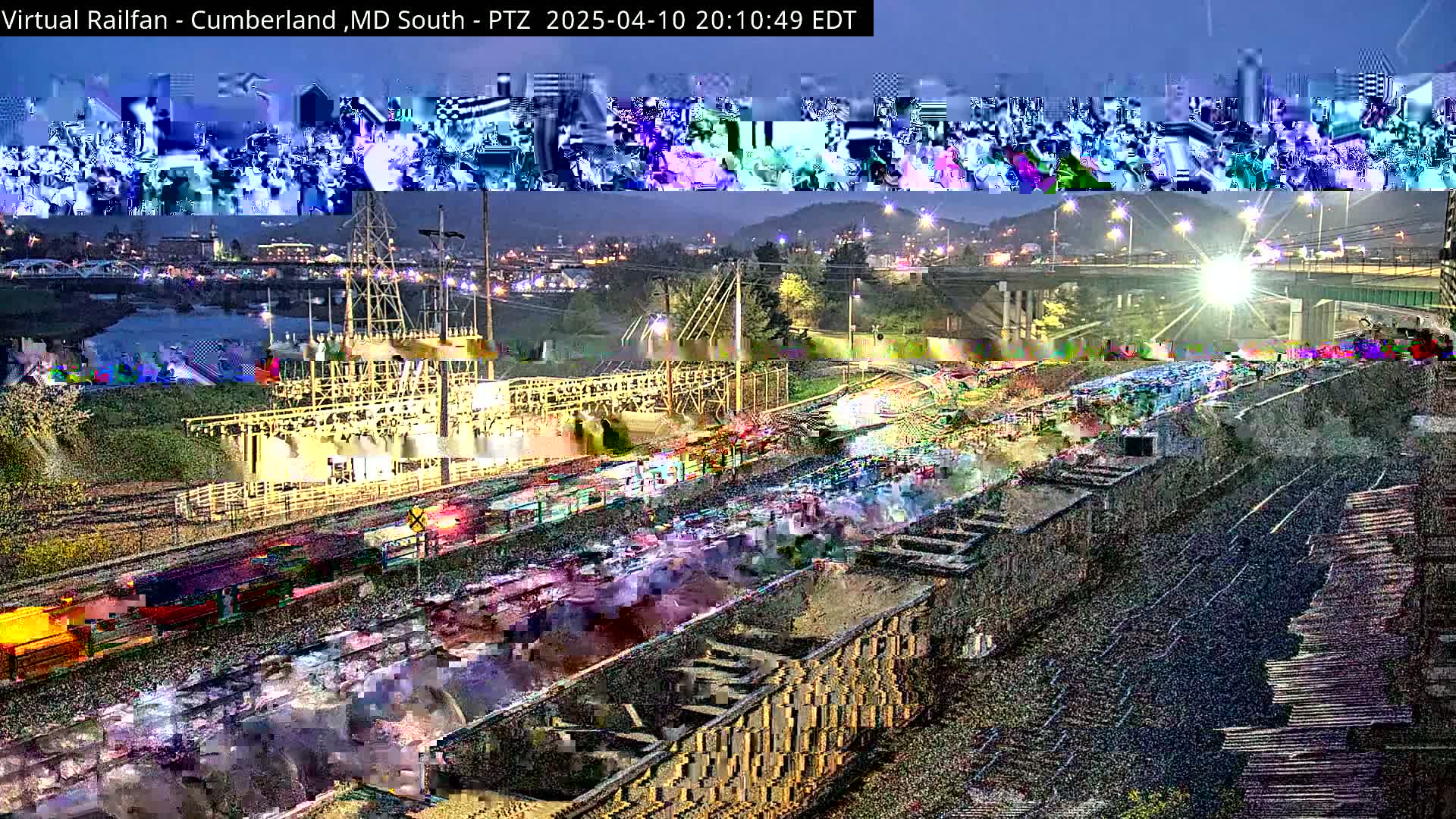 A nighttime view of a rail yard with numerous train cars and a bridge in the background under a twilight sky.