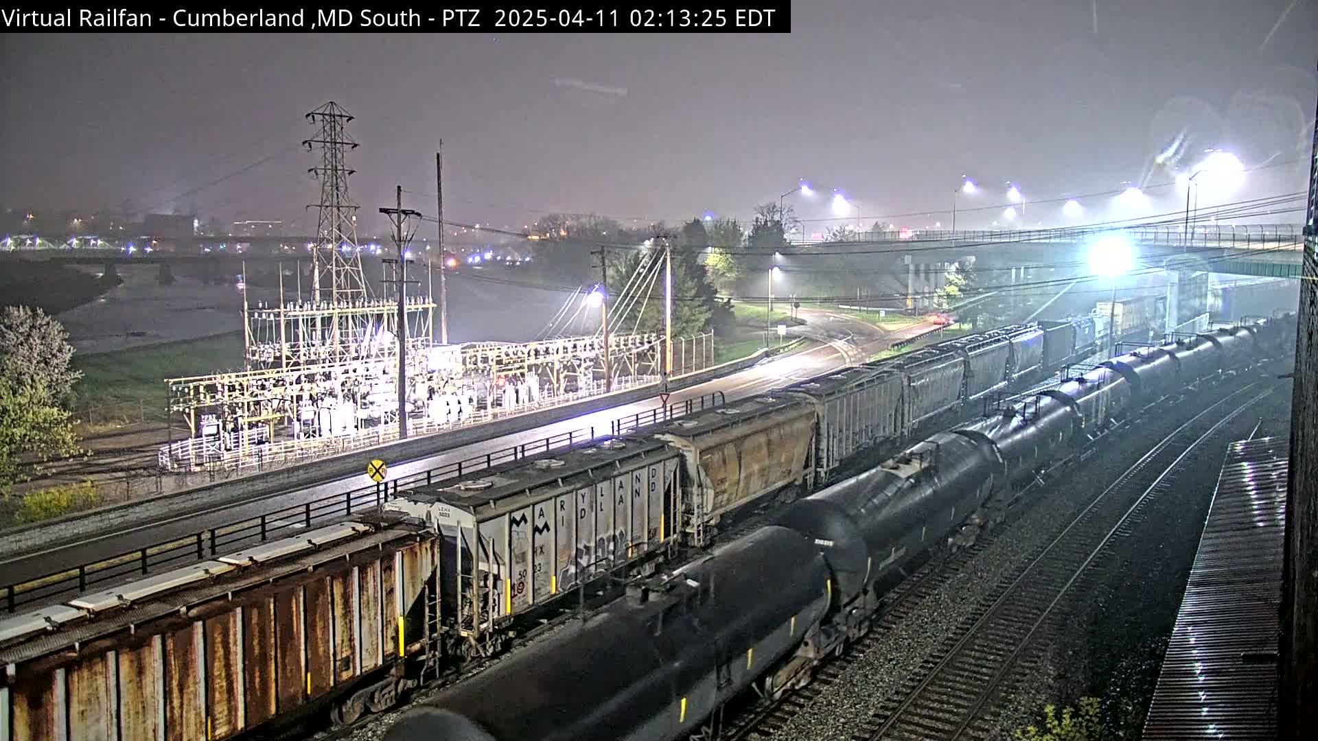 A long freight train, comprised of boxcars and tankers, is seen at night under hazy conditions near a power substation and a river.