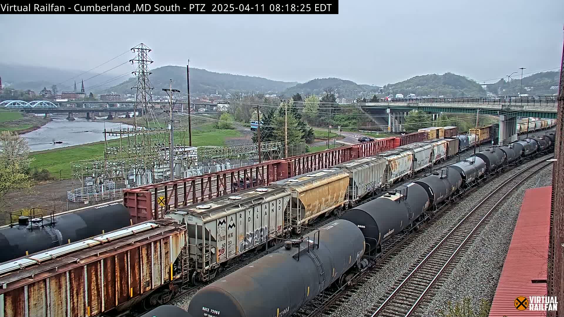 A long freight train, consisting of both boxcars and tanker cars, curves along a set of railroad tracks under an overcast sky, with a river and town visible in the background.