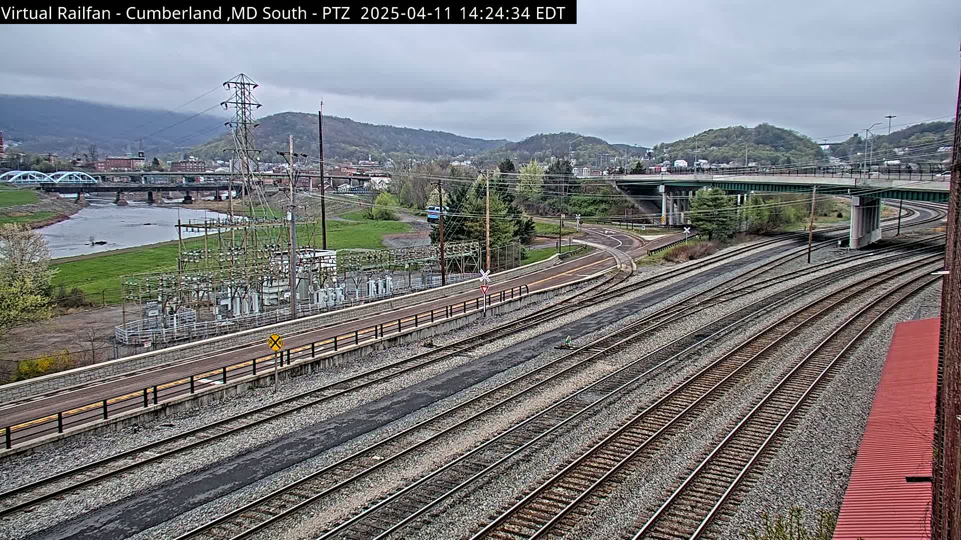 A sprawling railway yard with multiple tracks under an overcast sky, next to a river and a power substation, in a valley surrounded by hills.