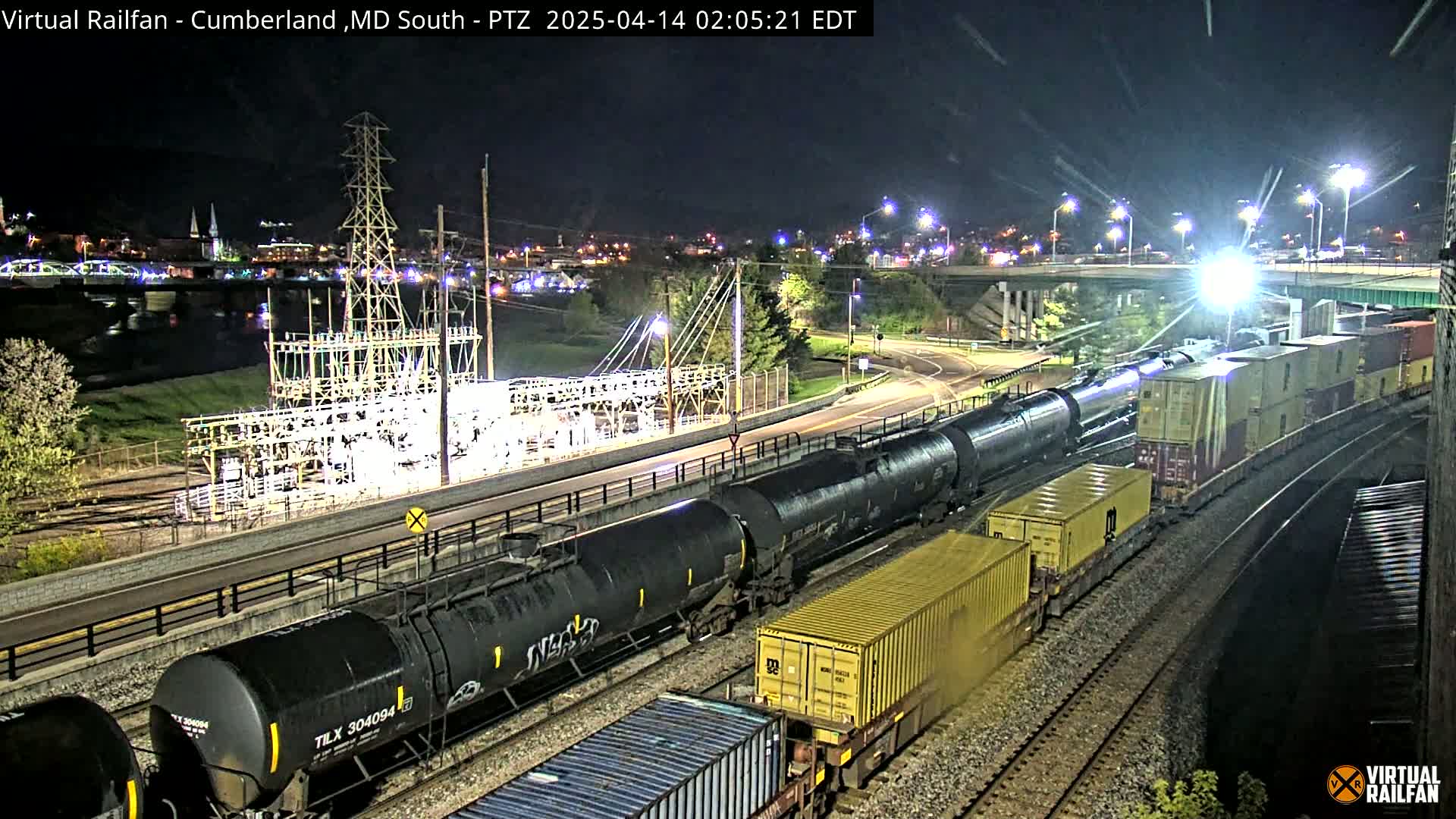 At night, a long freight train carrying tanker cars and shipping containers moves along railroad tracks near a power substation and a river, under well-lit conditions.