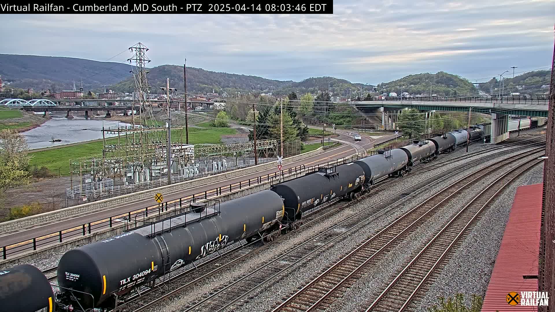 A long freight train of black tanker cars is seen on a set of train tracks next to a river, under an overcast sky, with a town and mountains in the background.