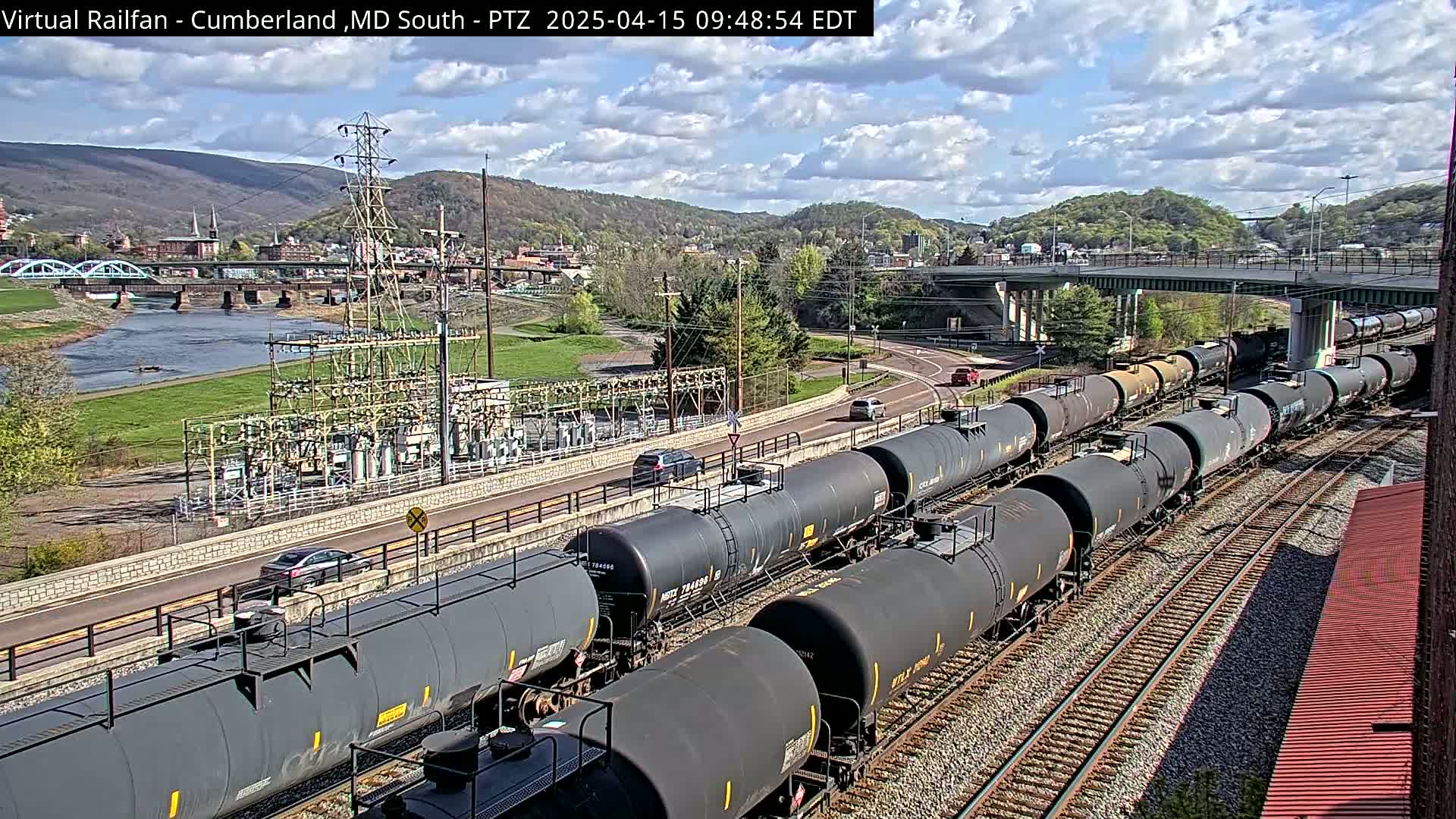 A long train of tanker cars sits on railroad tracks beside a river and road, under partly cloudy skies.