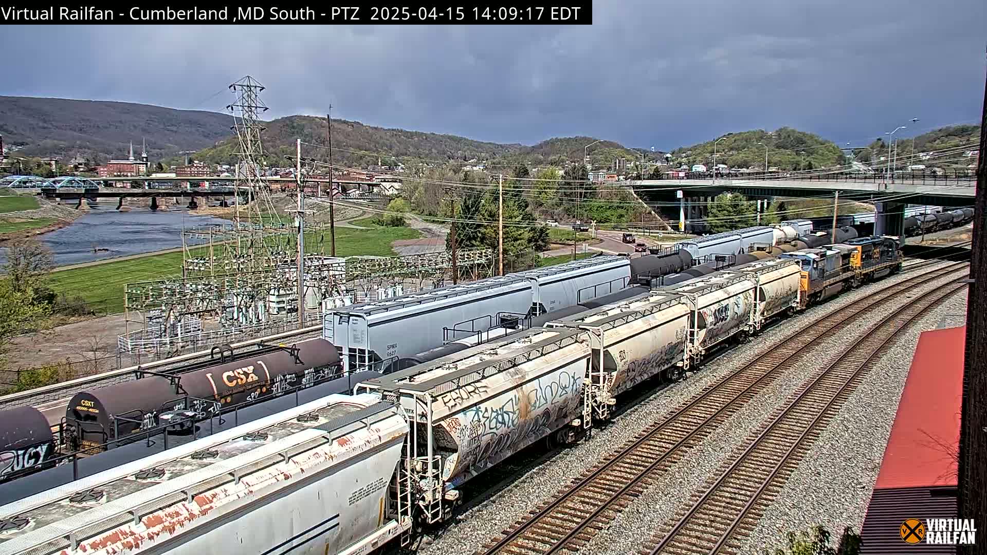 A long freight train, under partly cloudy skies, travels along train tracks near a river and a town nestled in a valley.
