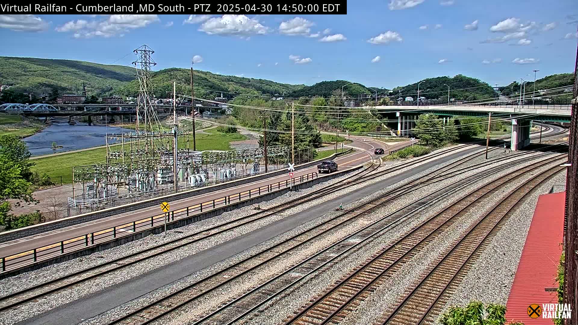A high-angle view shows a sunny day overlooking a river, a bridge, railroad tracks, an electrical substation, and a highway overpass, with a few cars visible on the roads.