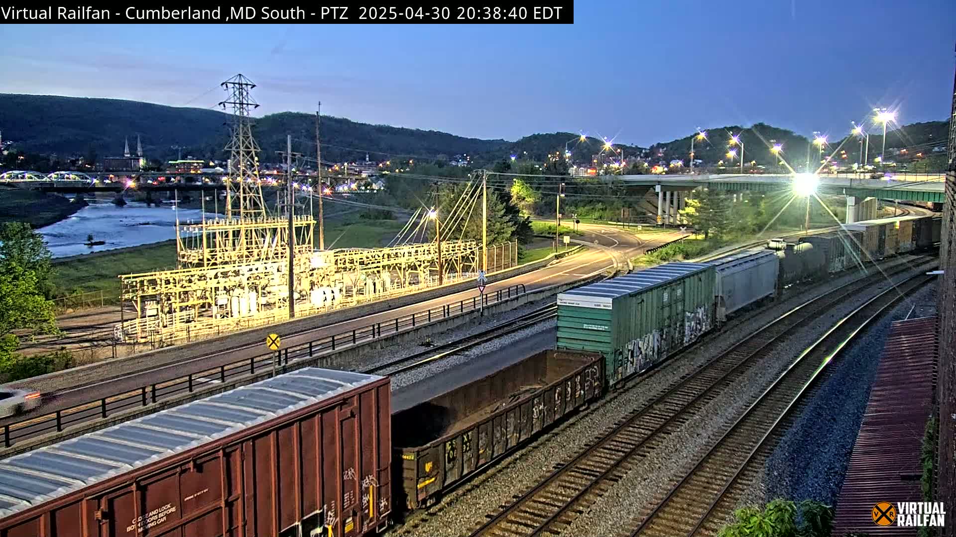 A nighttime view of a river, a town nestled against hills, a rail yard with several freight cars, and an electrical substation, all under a twilight sky.
