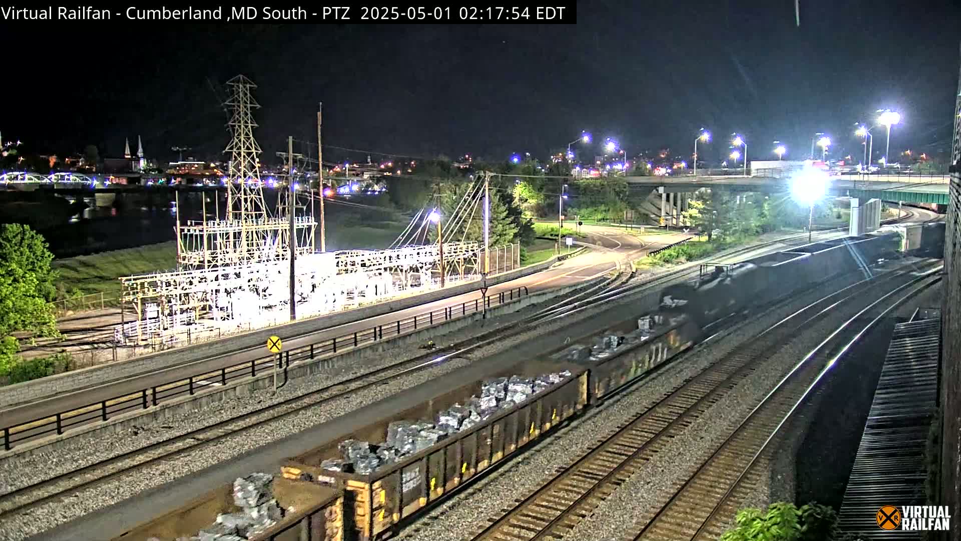 At night, a train carrying a cargo of what appears to be metal scraps is seen on railway tracks next to a power substation near a river and a highway overpass.
