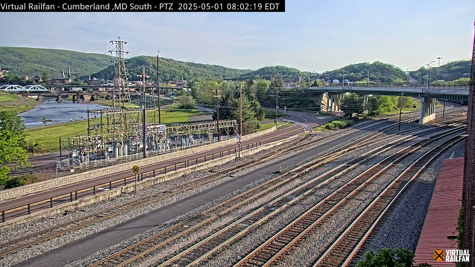 A sunny view of a river, railroad tracks, a road, and an electrical substation, set against a backdrop of green hills.
