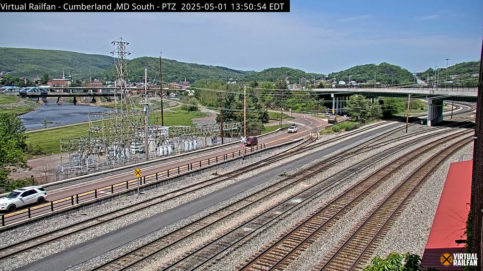 A high-angle view shows a sunny day overlooking railroad tracks, a river, a bridge, and a town nestled in a valley surrounded by green hills.
