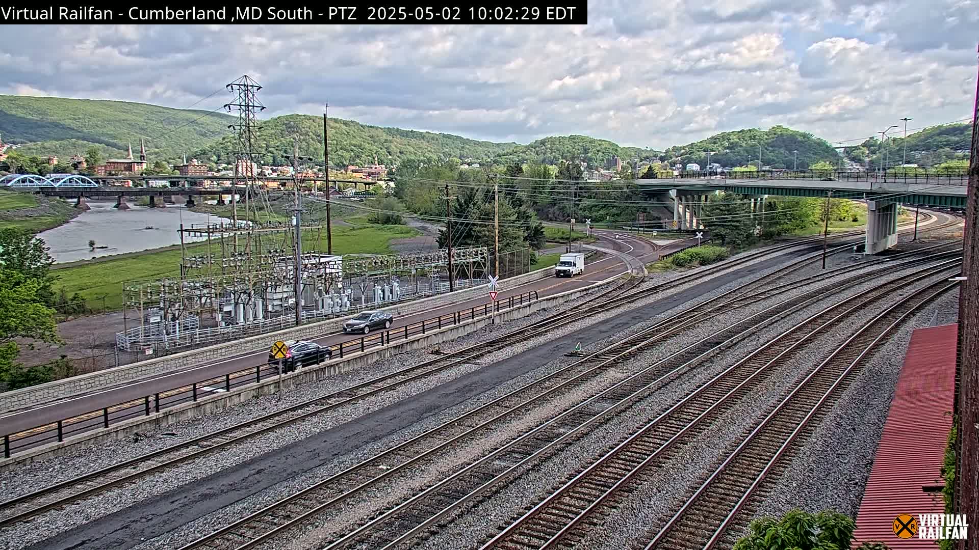 A view of a river, a town, and multiple sets of train tracks under a partly cloudy sky, with several vehicles visible on a road near the tracks and a power substation.