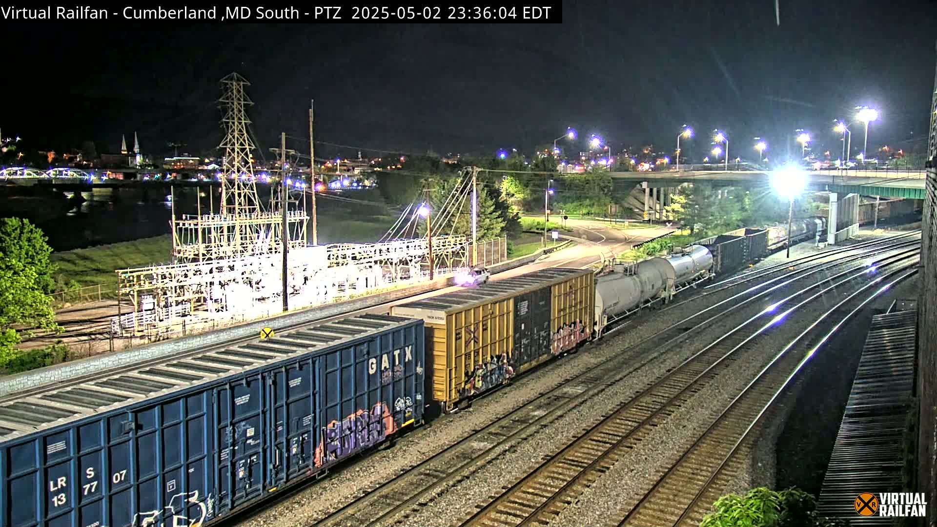 A nighttime view of a train passing by a power substation and a river, under a clear sky.