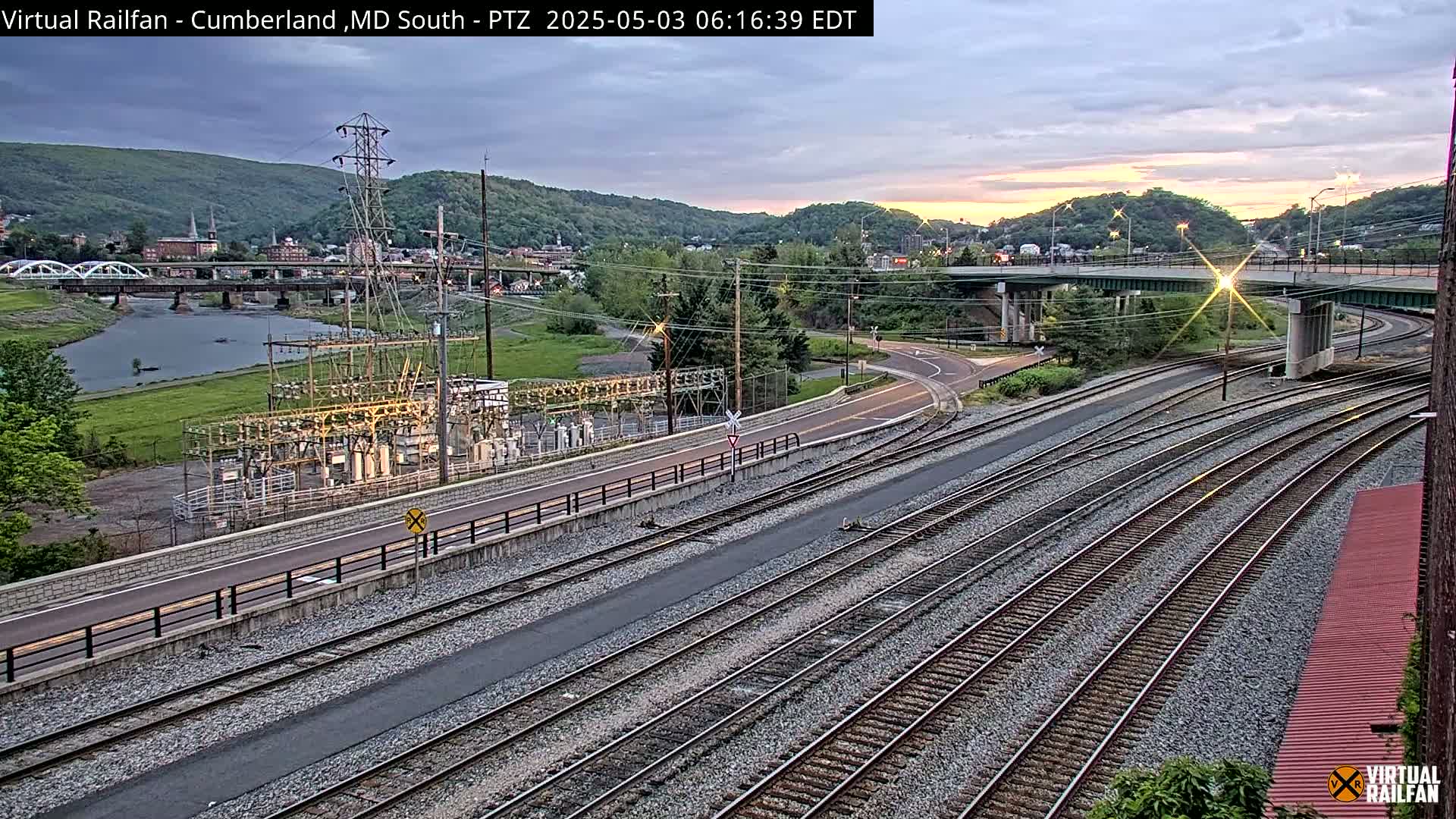 A twilight view of a river, a town nestled in hills, a power substation, and multiple sets of railroad tracks converging near a roadway intersection.