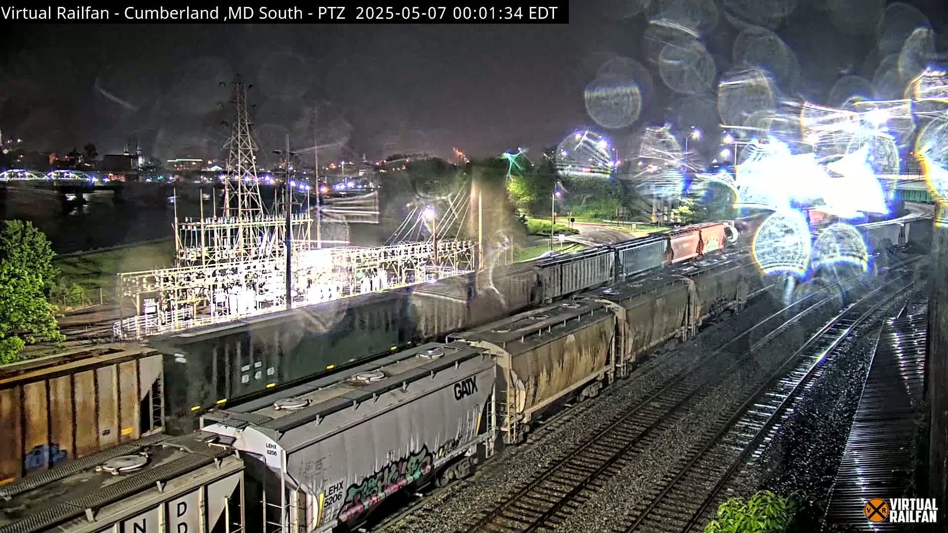 A long freight train is seen passing by an electrical substation at night in the rain.