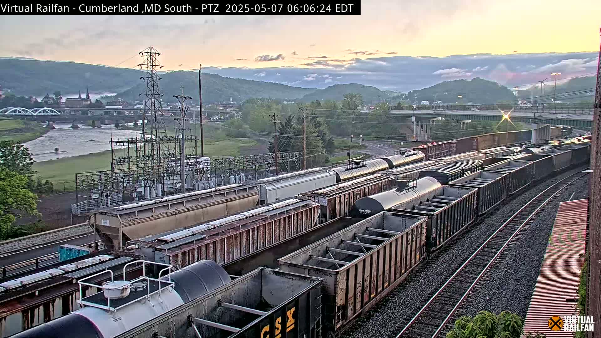 A long freight train sits on railroad tracks next to a river, with a town and mountains visible in the background under a sunrise sky.