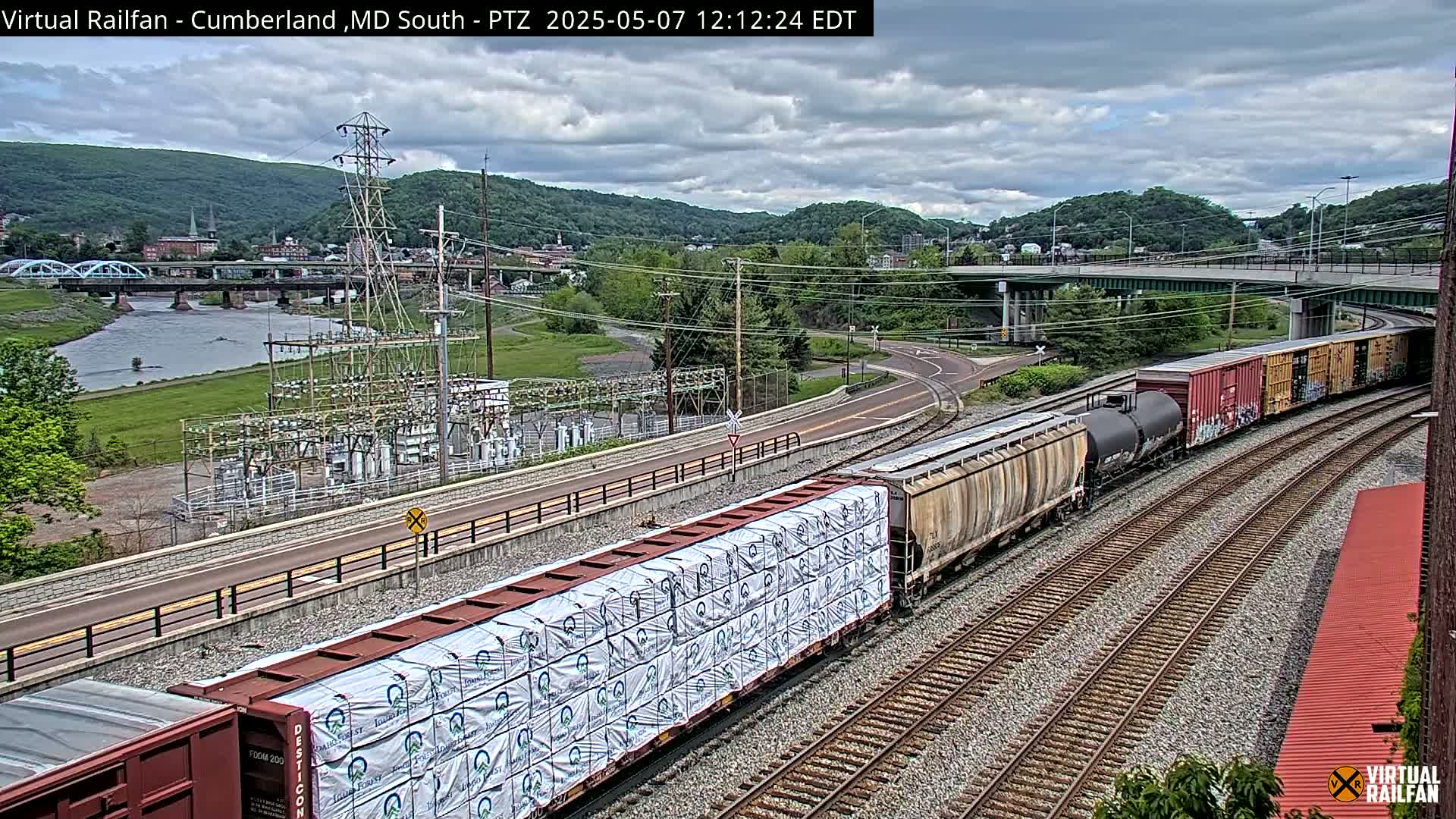 A freight train carrying lumber passes by a river, a bridge, and an electrical substation under a partly cloudy sky.