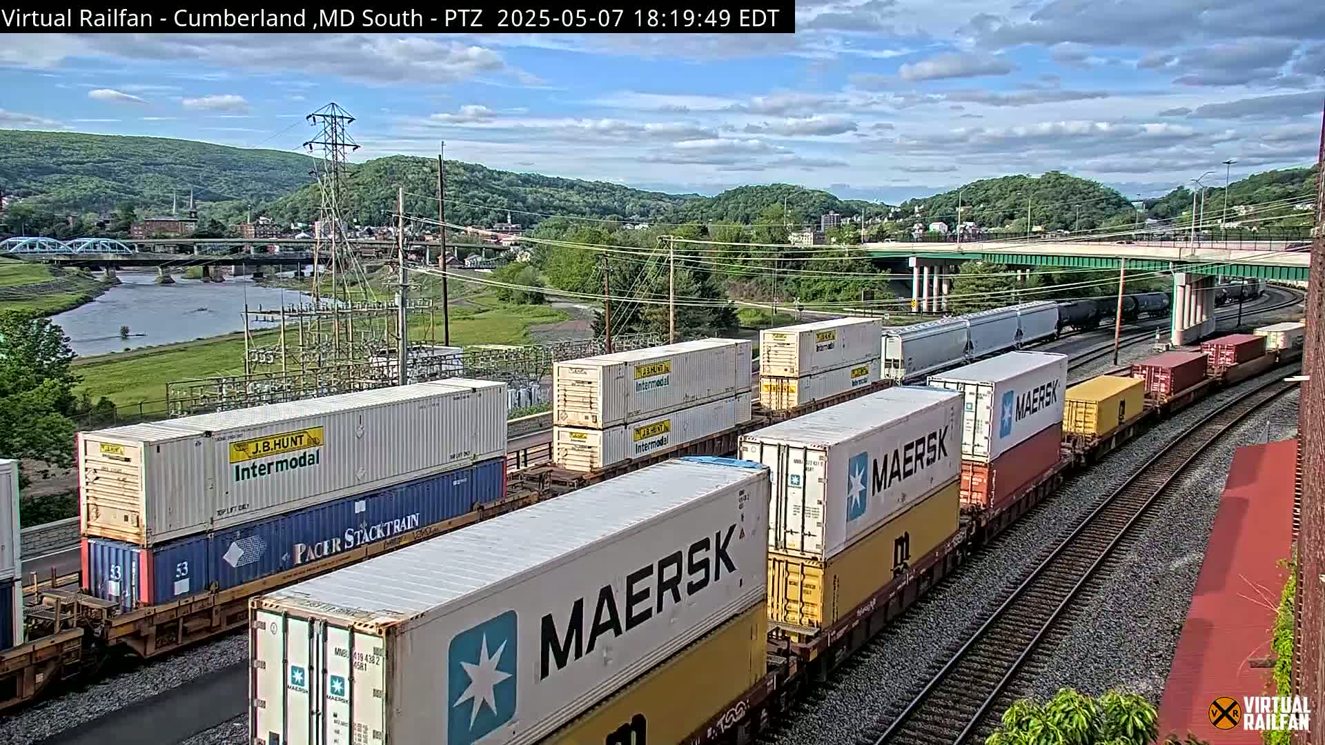 A long freight train carrying numerous shipping containers is seen on a rail line next to a river, under a partly cloudy sky.