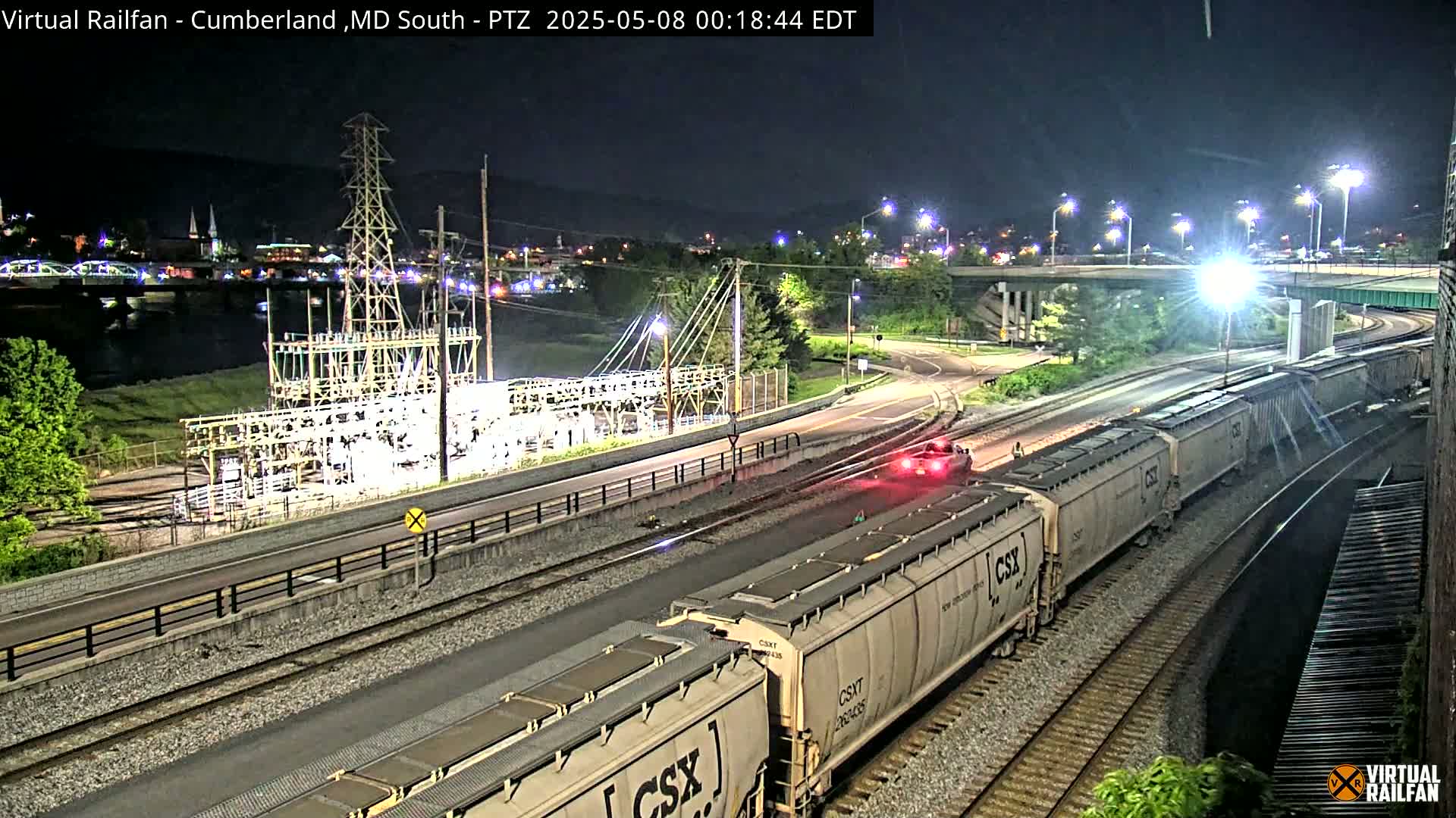 At night, a long freight train is moving along railroad tracks near a highway and a power substation, with a river and city lights in the background.