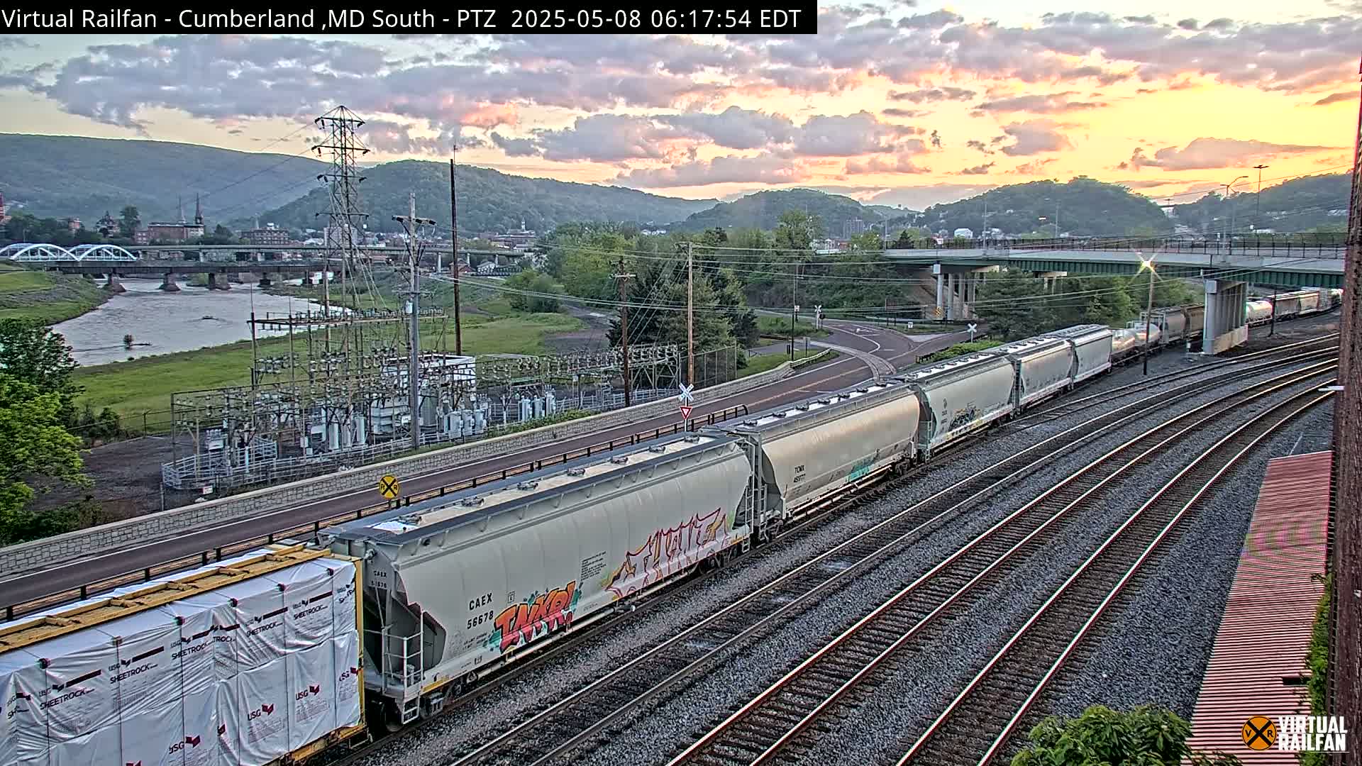 A long freight train passes under a bridge at sunset, next to a river and power station, in a valley.
