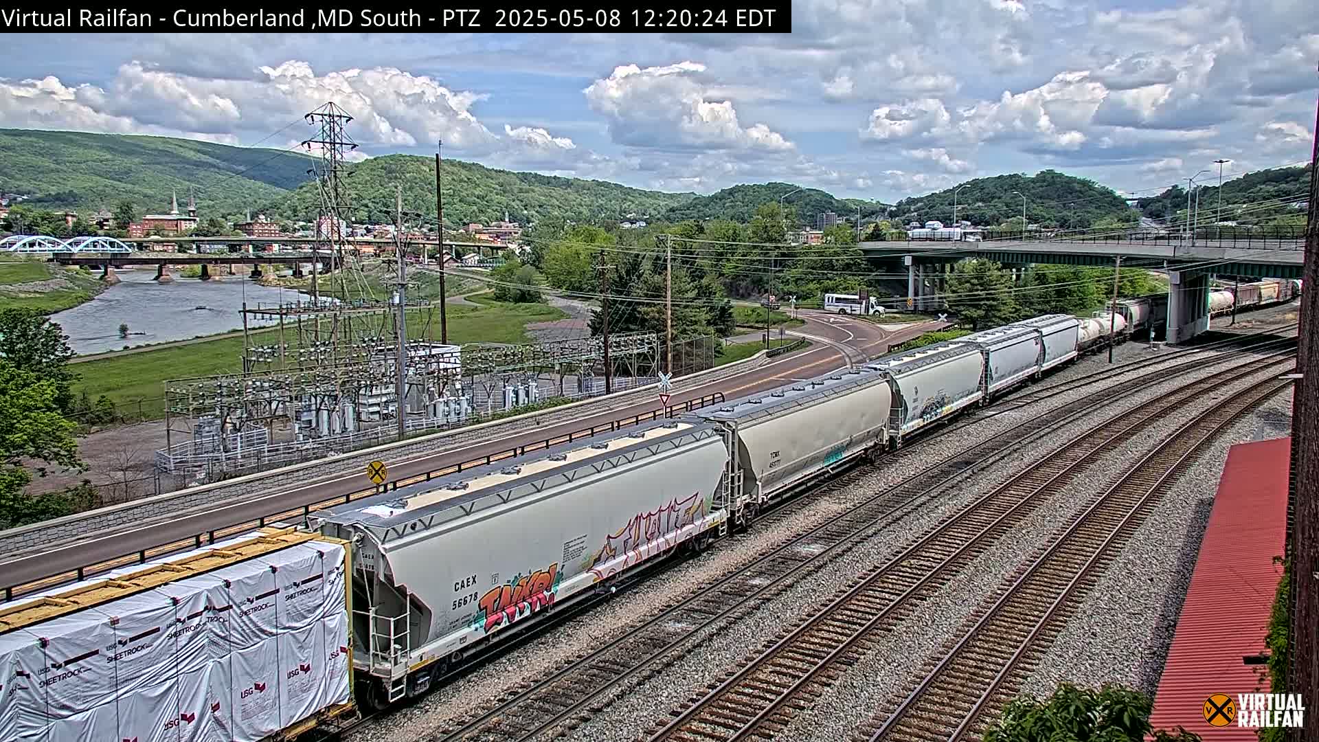 A long freight train passes alongside a river, under a bridge and past an electrical substation, on a partly cloudy day.