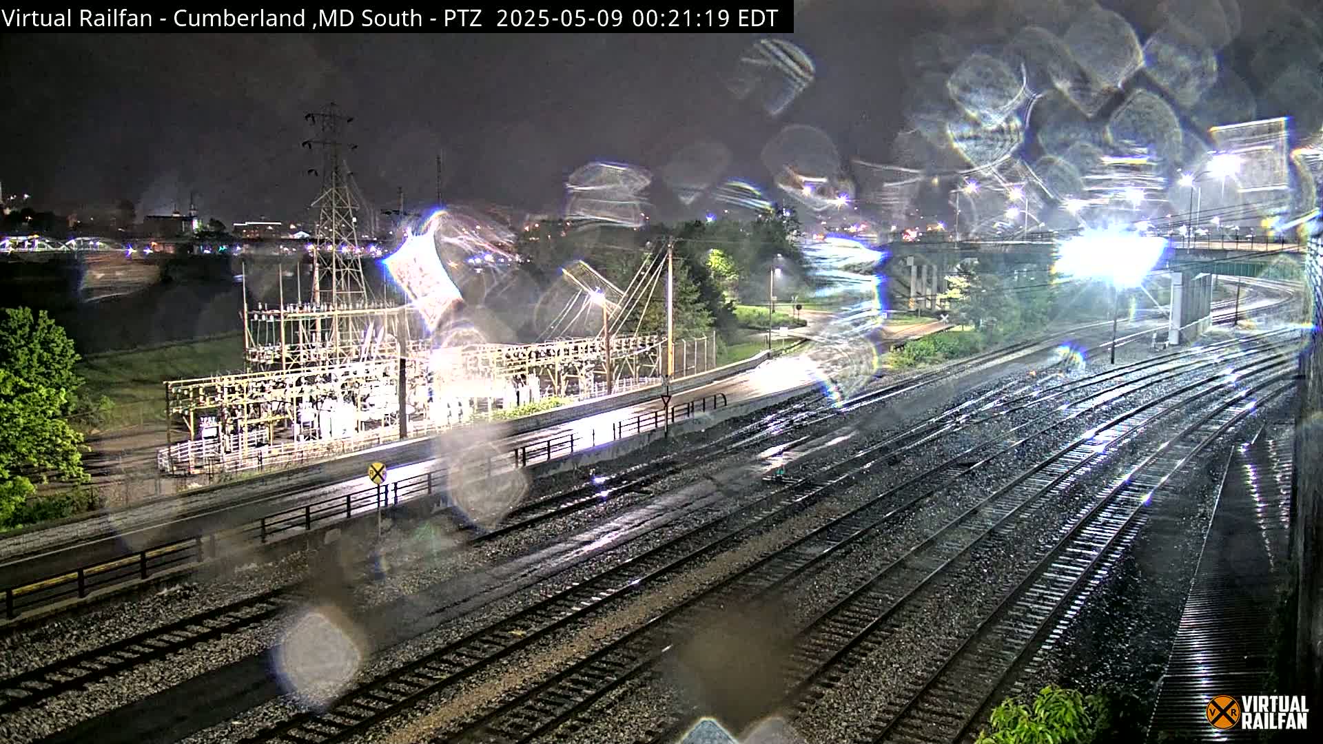 A nighttime view of rain-slicked railroad tracks alongside an electrical substation, under a dark, rainy sky.