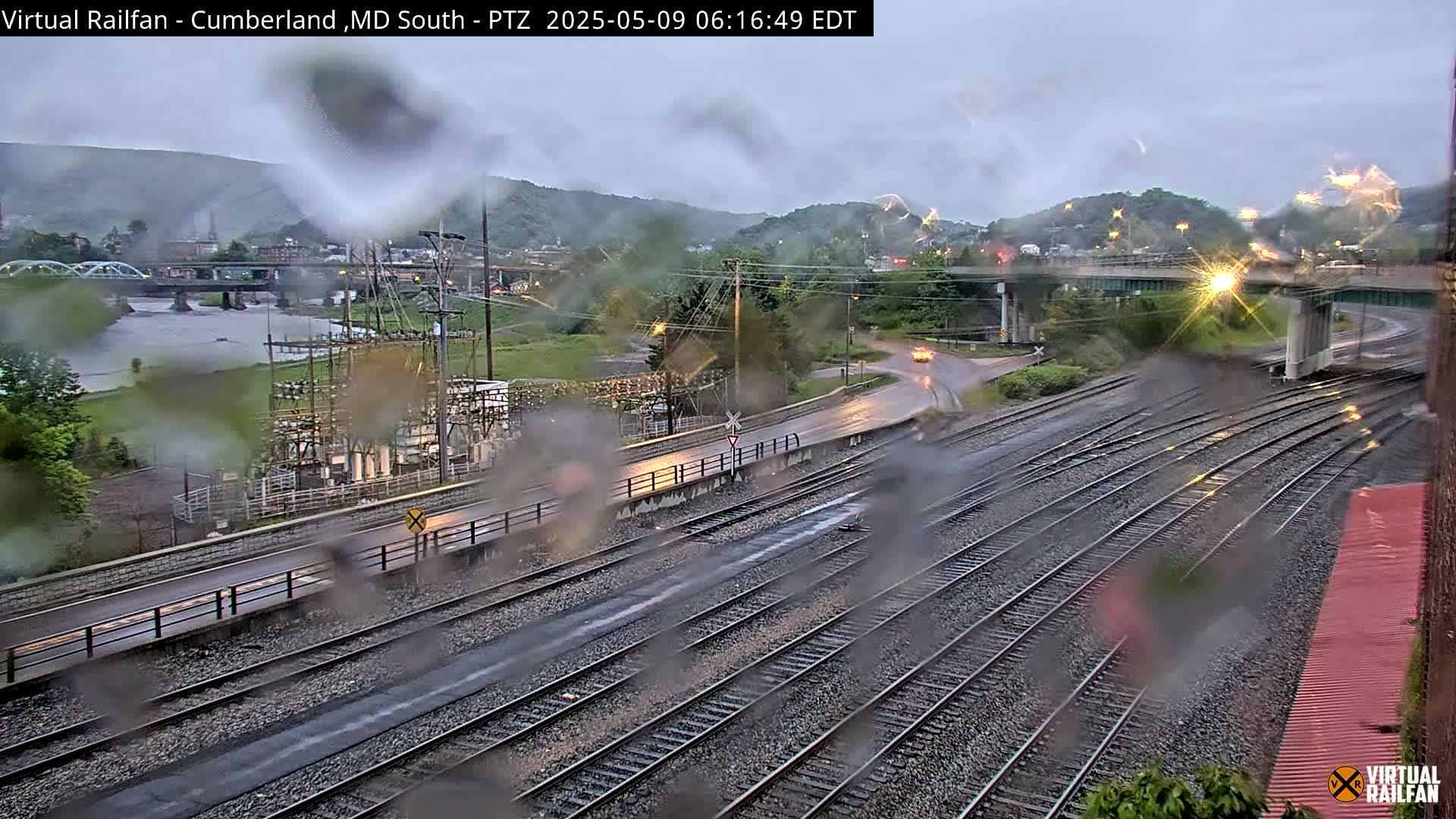 A rain-slicked view of railroad tracks and roads intersecting near a river and bridge, with hills in the background under an overcast sky.