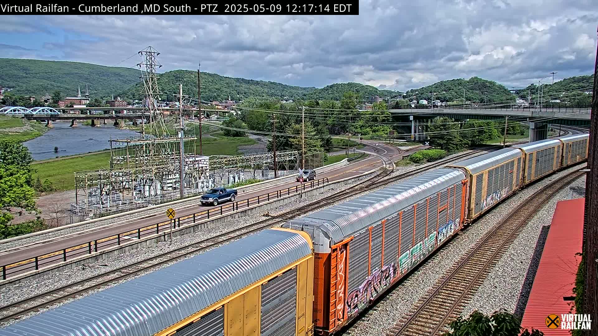 A long freight train passes under a highway overpass on a partly cloudy day, near a river and a power substation.