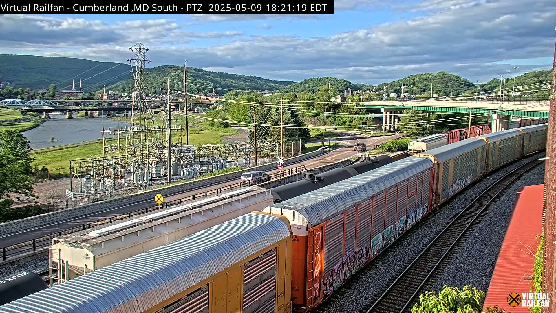 A long freight train curves along railroad tracks next to a river, under a partly cloudy sky, with a bridge and power station visible in the background.