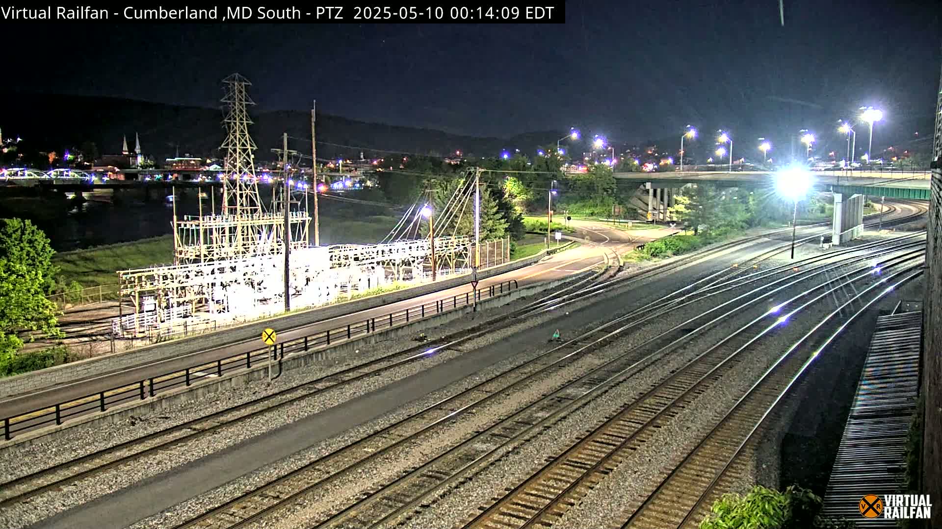 A nighttime view of railroad tracks, a roadway, and an electrical substation, with a river and town in the background under clear skies.
