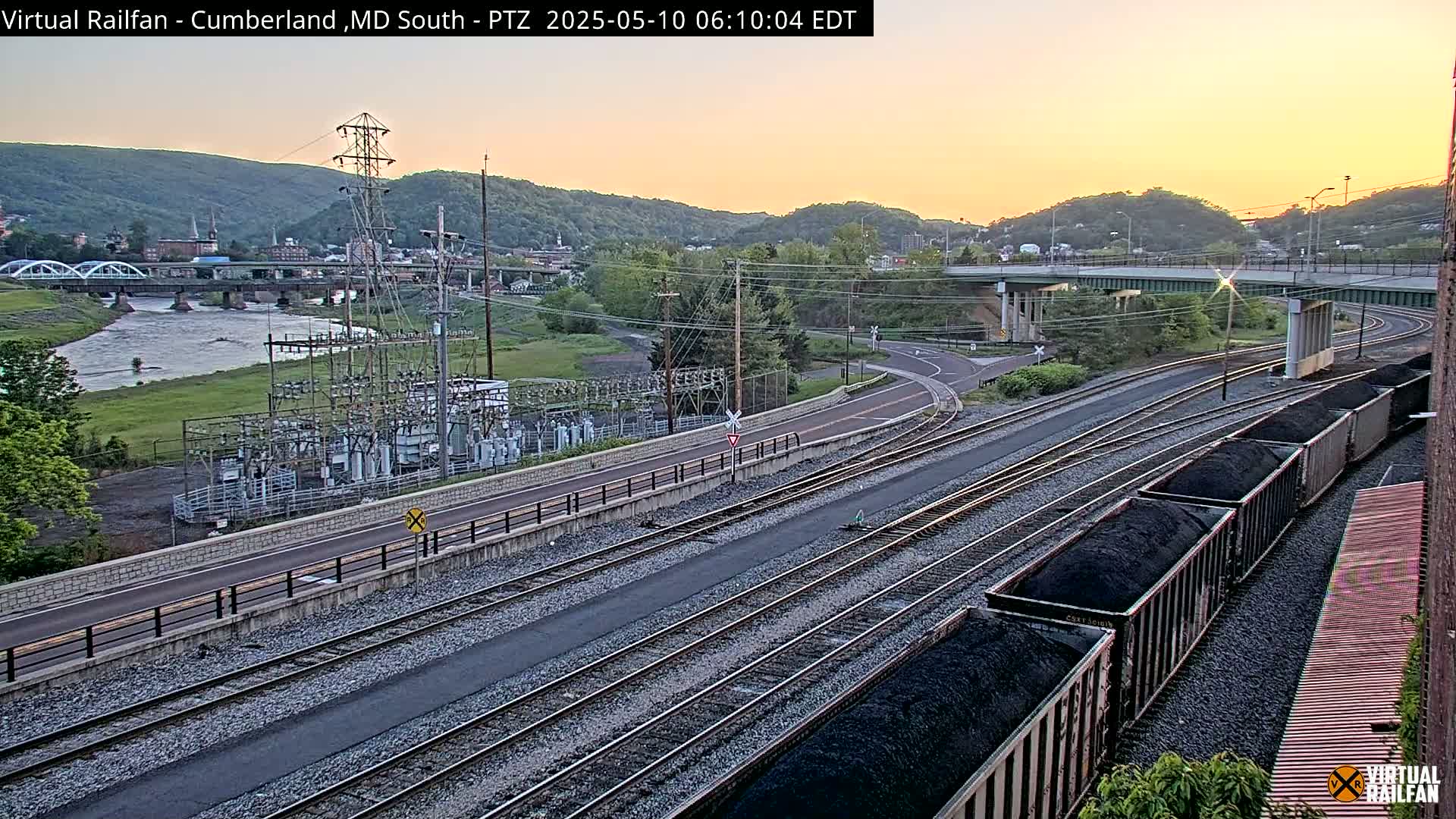 A high-angle, long shot shows a river, a town, a power substation, railroad tracks with coal cars, and a highway overpass during a sunrise with clear skies.
