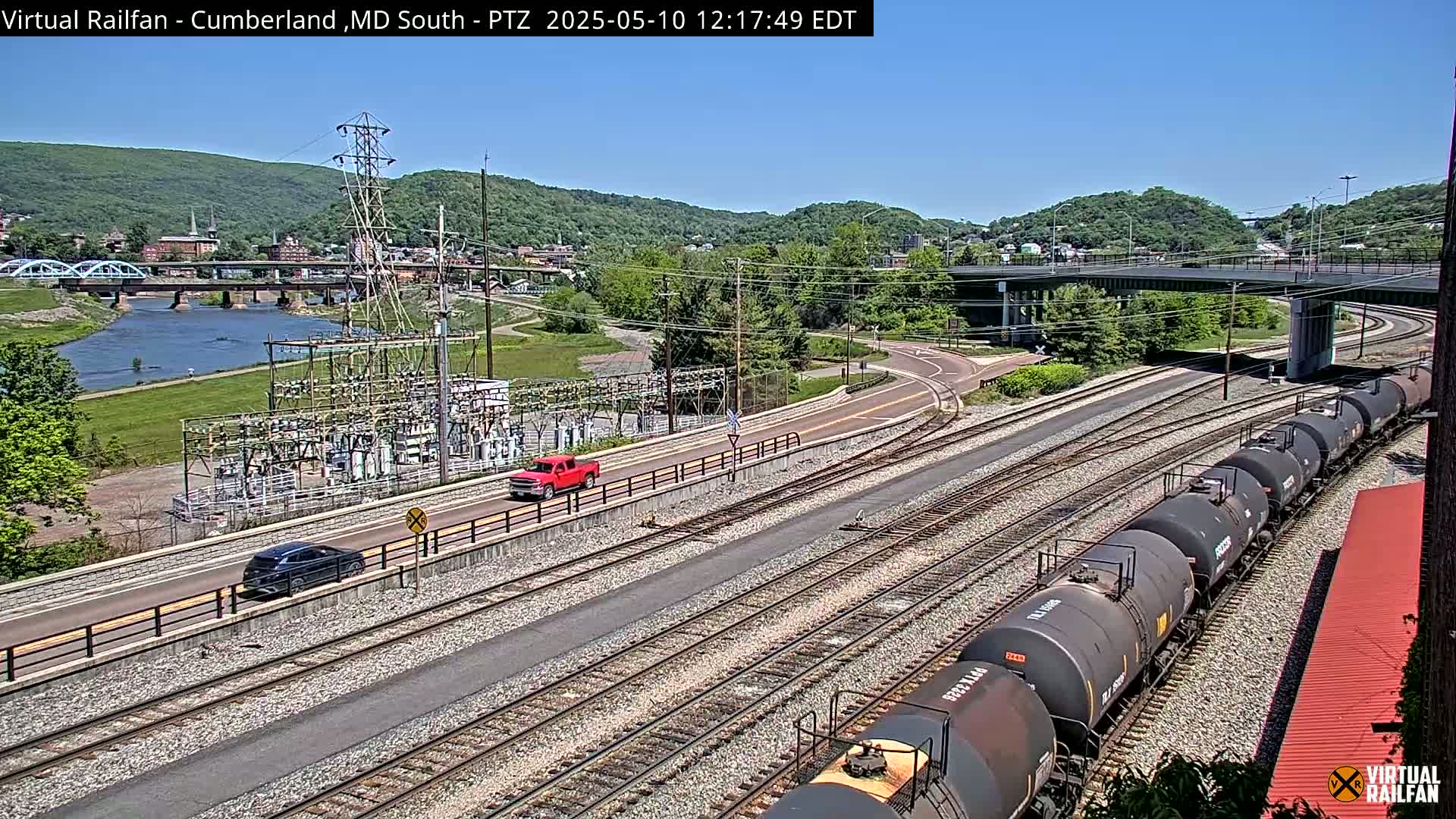 A sunny day overlooking a river, train tracks with tanker cars, roads with vehicles, and a power substation, all situated beside a town nestled in a valley.