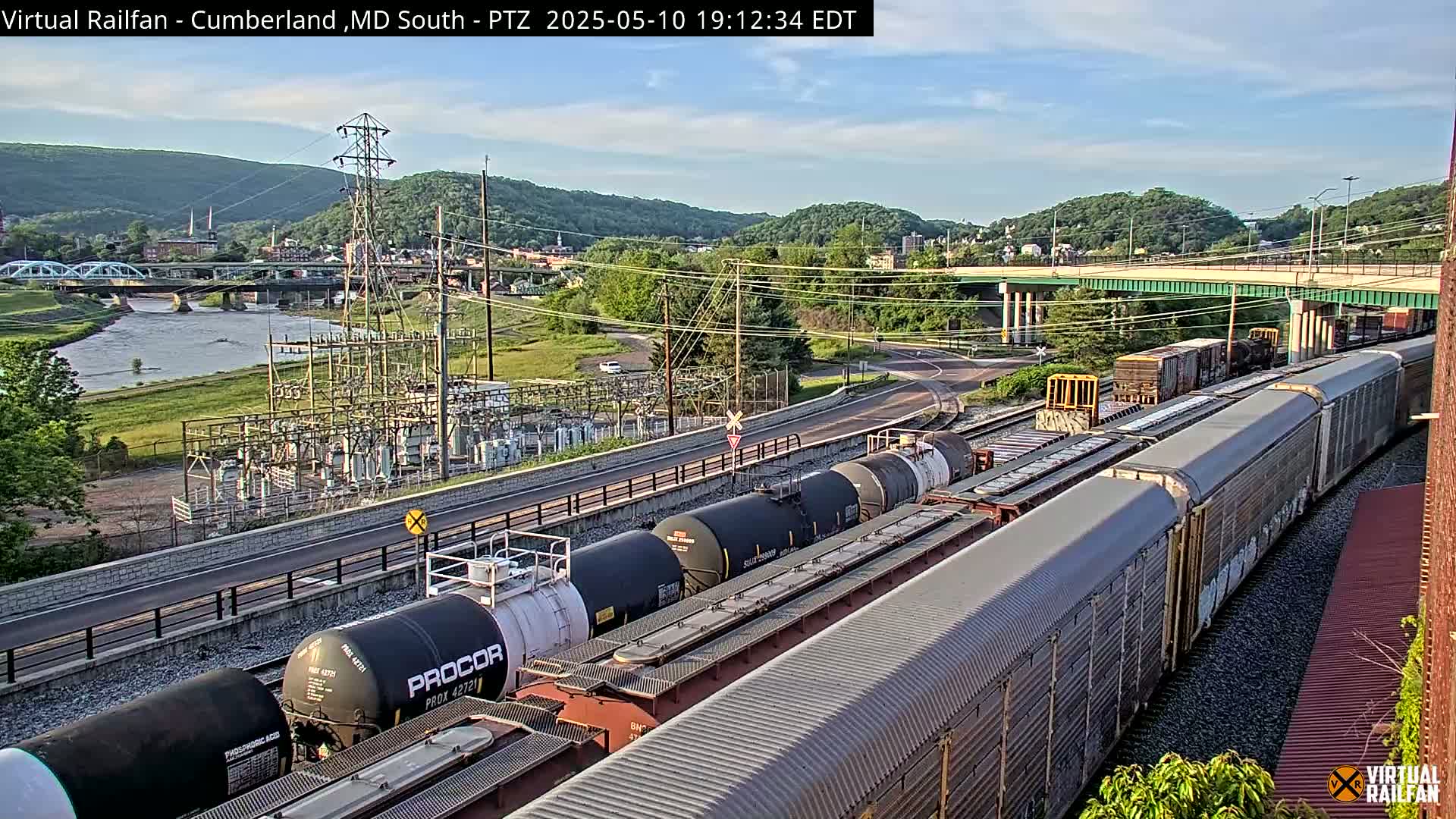 A long freight train carrying tanker cars and boxcars sits on tracks beside a river under a partly cloudy sky, with a bridge and power station visible in the distance.