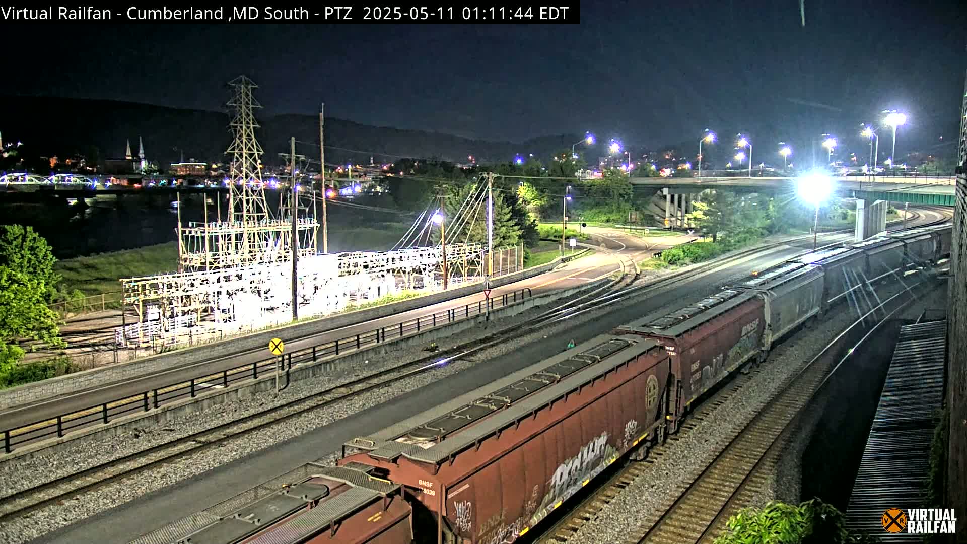 At night, a long freight train moves along railroad tracks near a river, passing an electrical substation and a road underpass, with a town visible in the background.