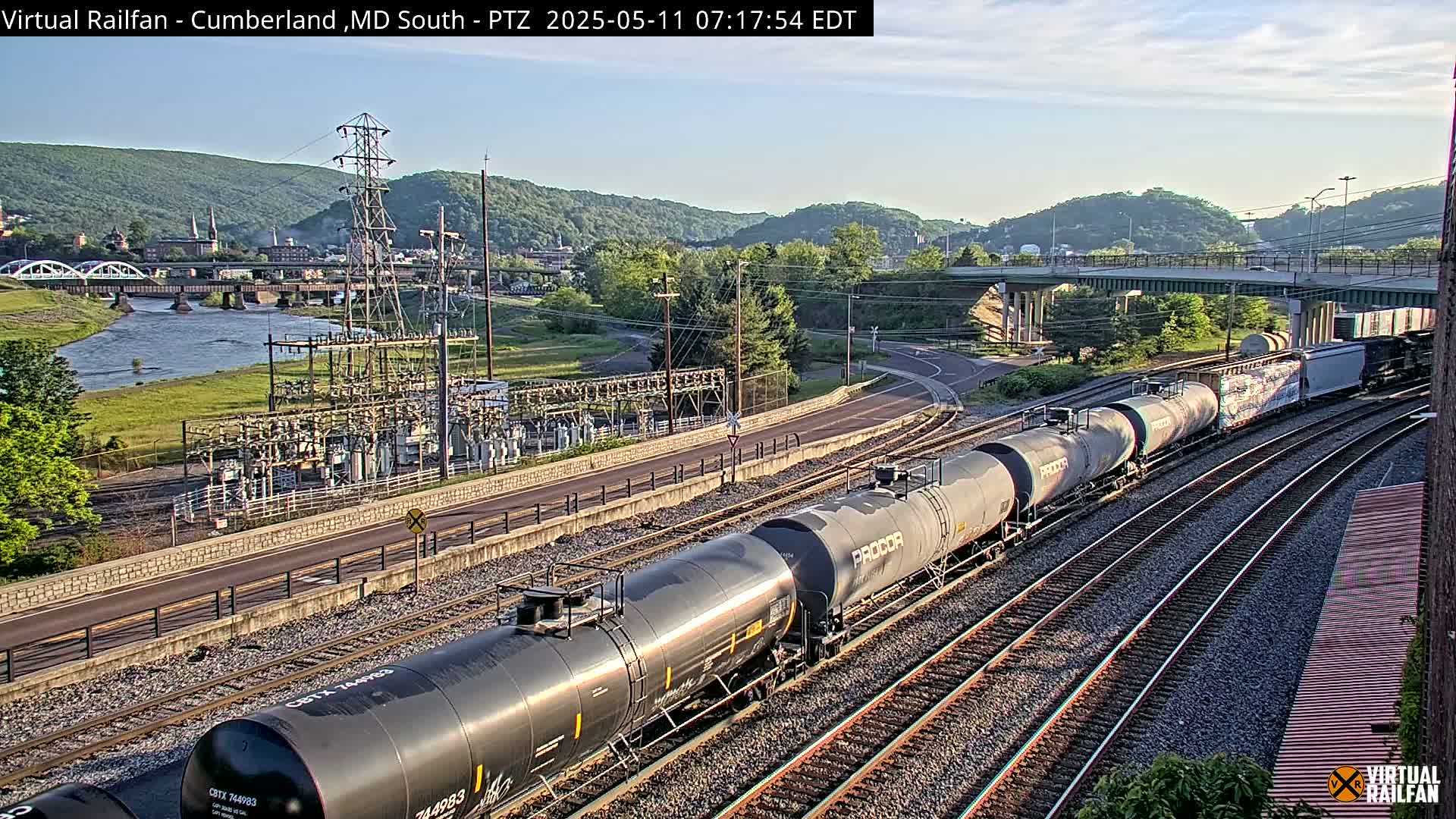 A long freight train of tanker cars is visible on a set of railway tracks, with a river, bridges, and a power substation in the background under a clear sky.