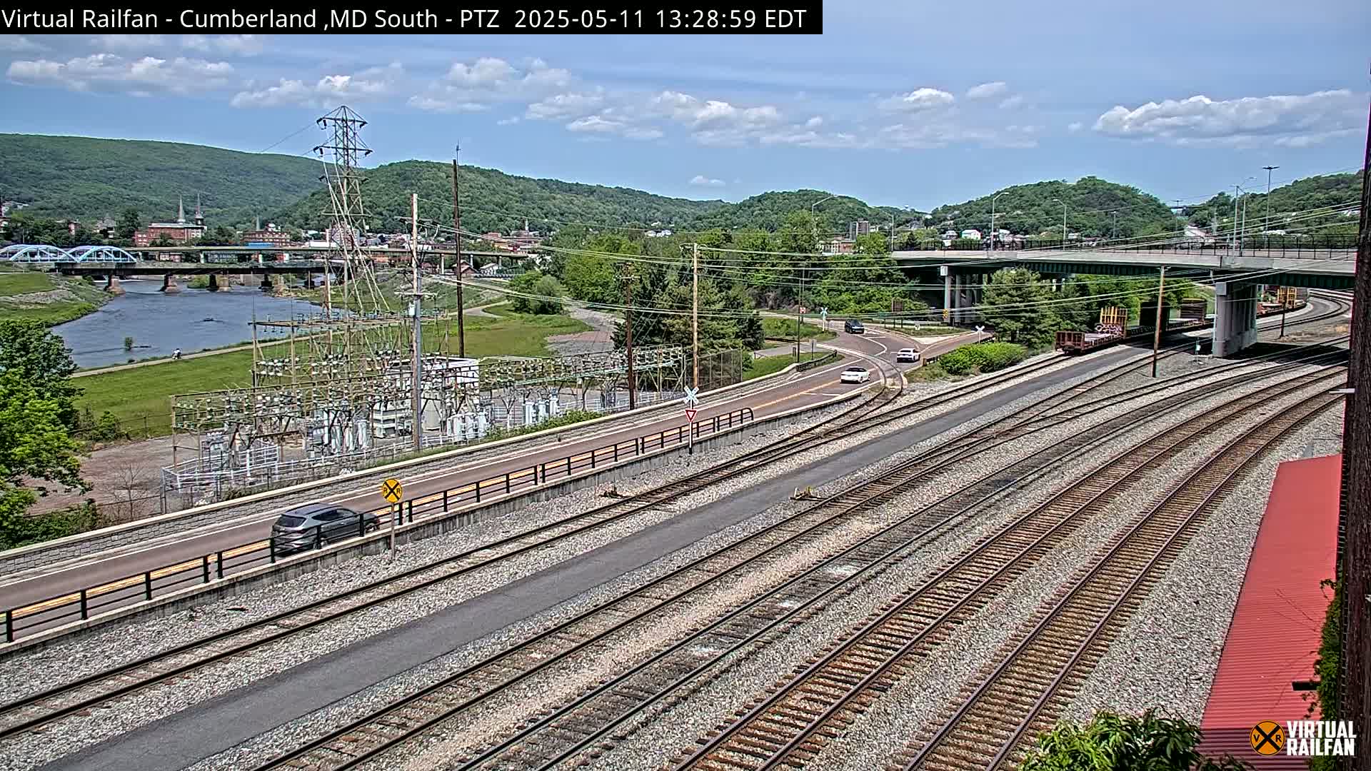 A high-angle view of a partly cloudy day showing railroad tracks, roads, a river, a bridge, and an electrical substation next to green hills.