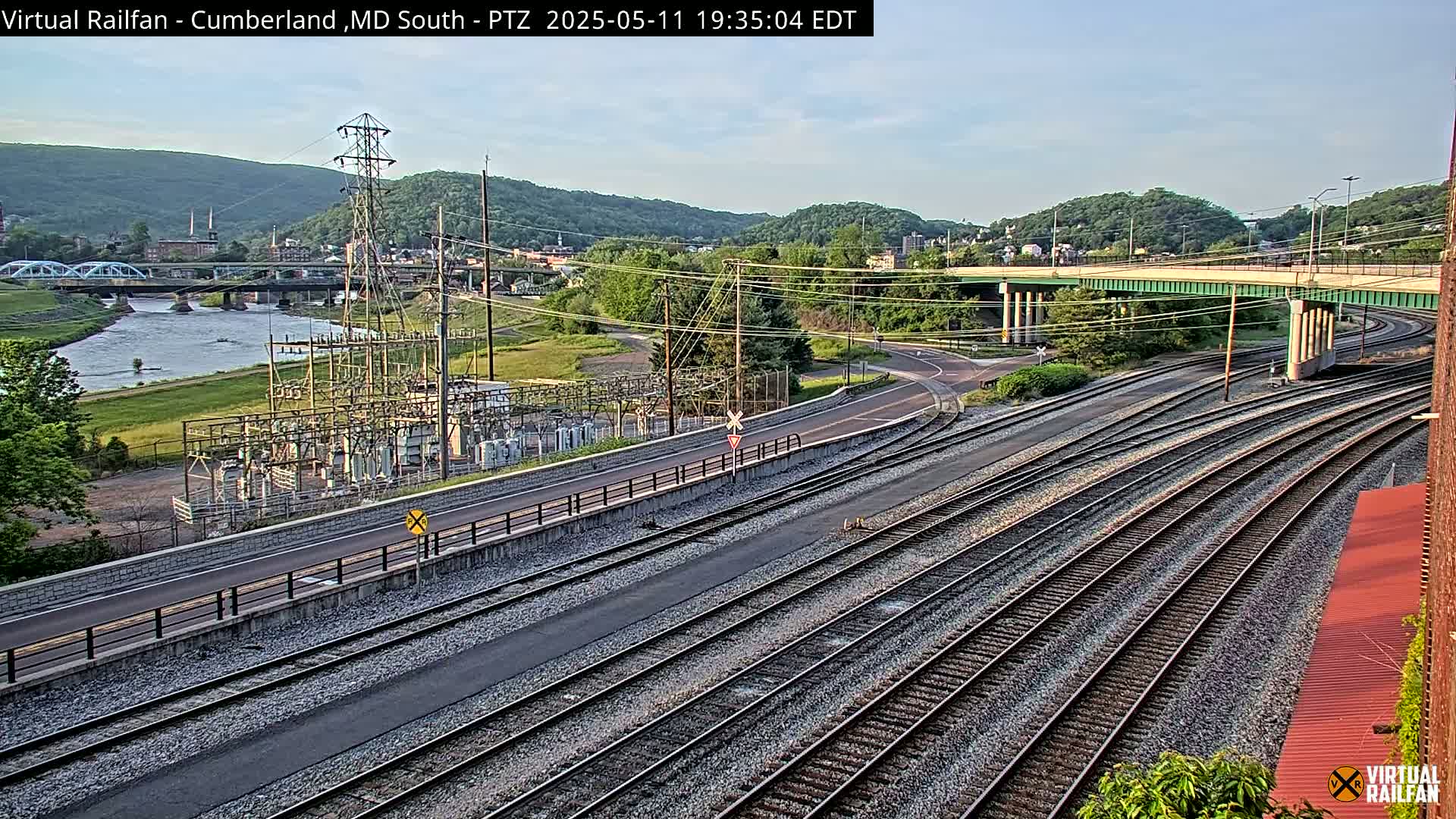 A view of railroad tracks and a road curving alongside a river under a partly cloudy sky, with a power substation and a town nestled in the background against a hillside.