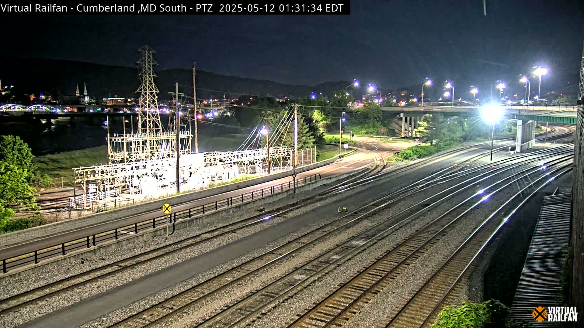 A nighttime view of several parallel train tracks next to a road and an electrical substation, with a river and city lights in the background.