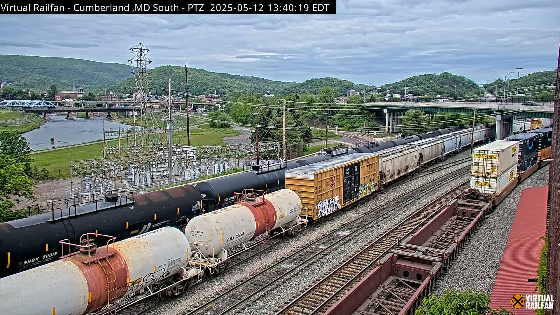 A long freight train, including tanker cars and shipping containers, sits on railroad tracks under an overcast sky, with a river and town visible in the background.