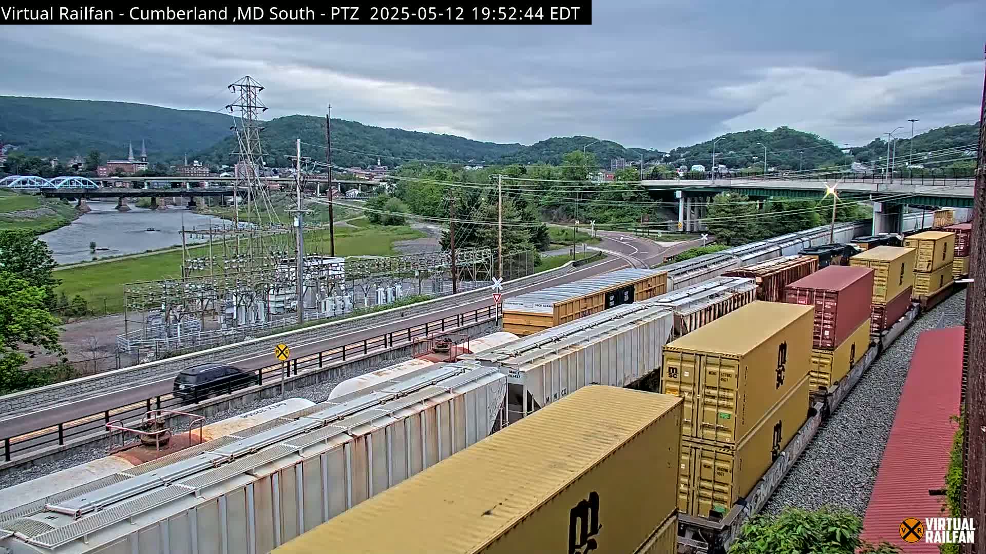 A long freight train with yellow and red shipping containers is visible, situated near a river, a bridge, and a power substation, under a cloudy sky.