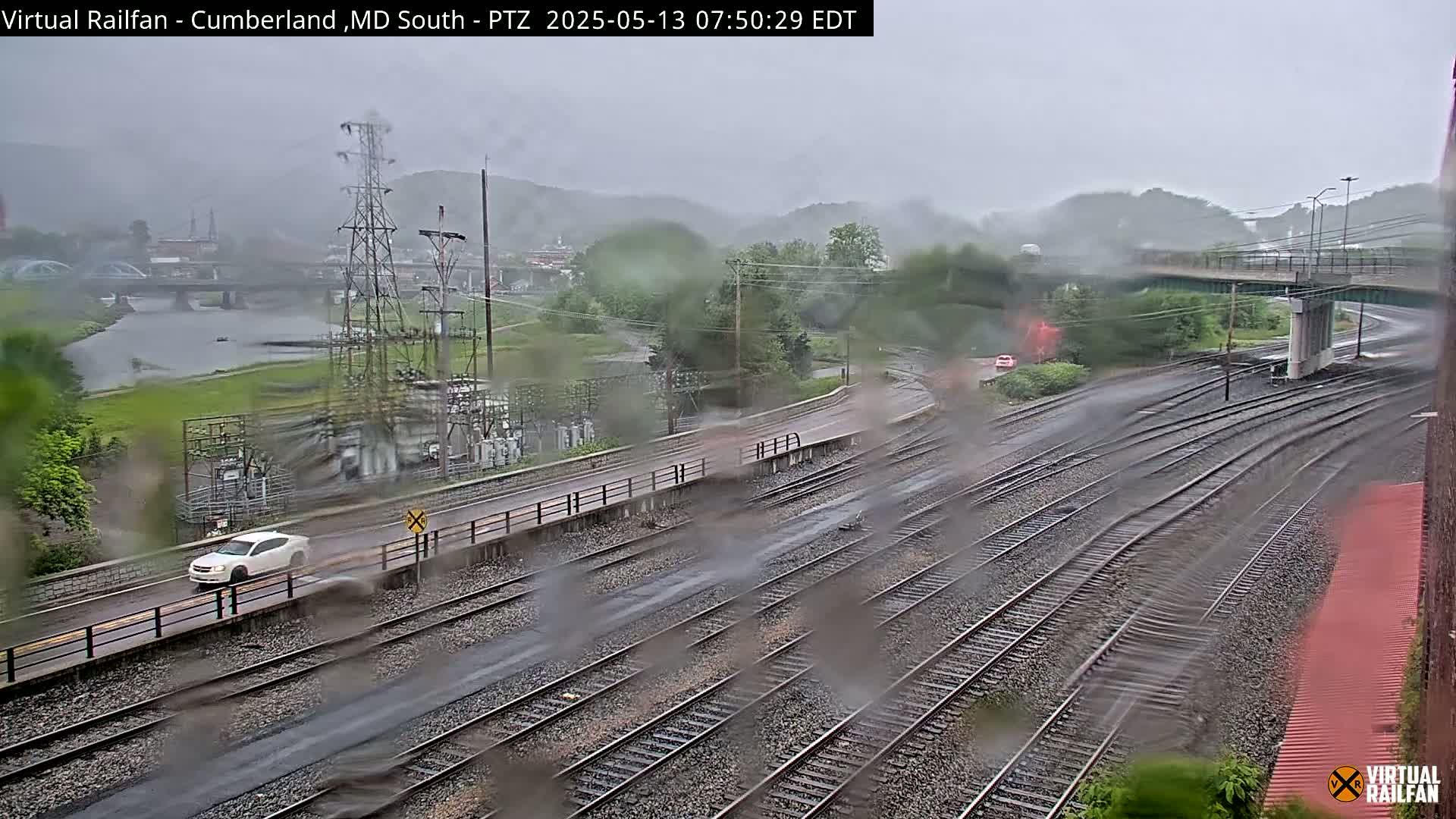 A rainy view of a railroad yard and a river, with a car driving on a road next to the tracks, and hills in the background.