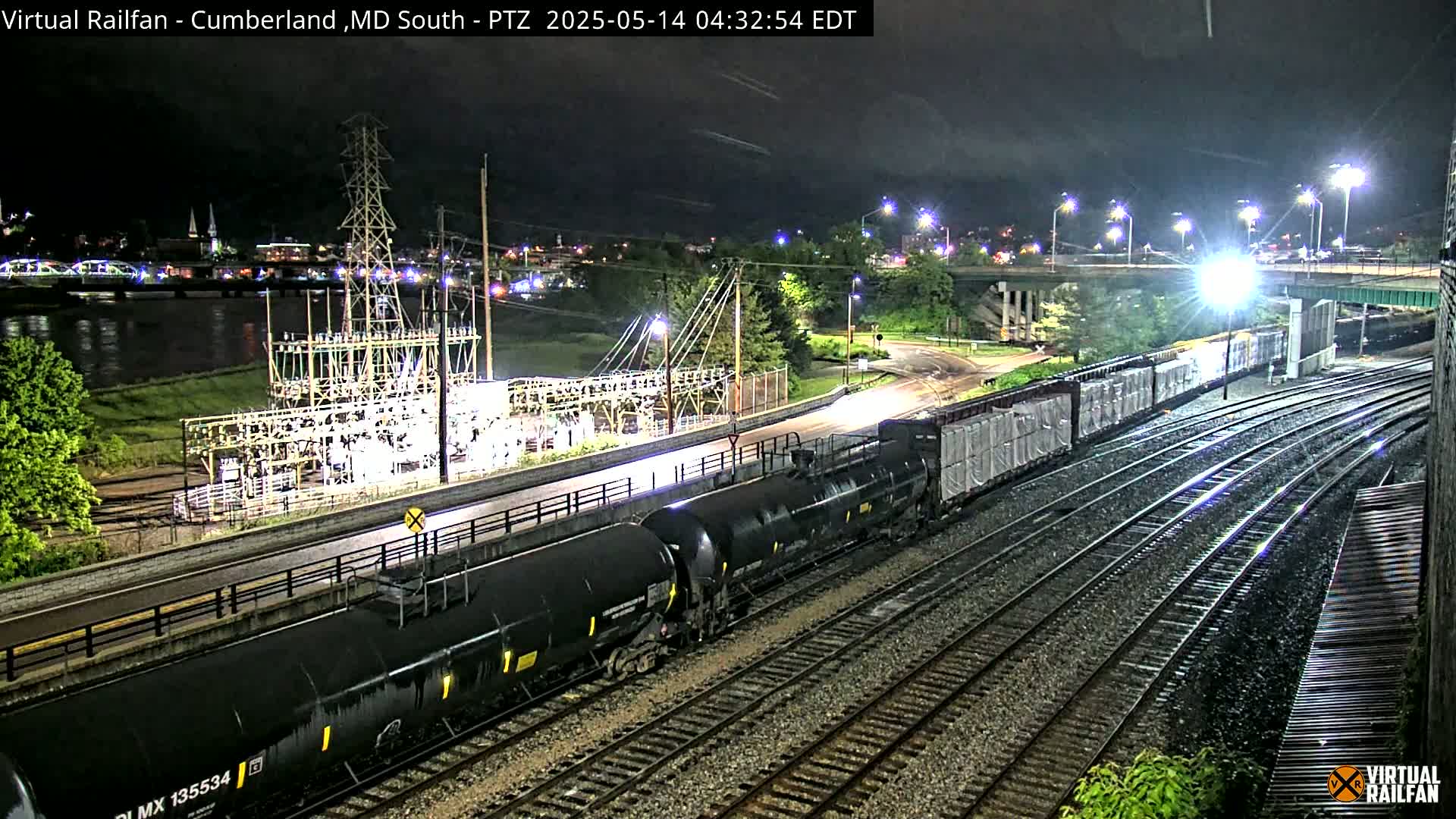 A long freight train passes at night near a river and power station under partly cloudy skies.